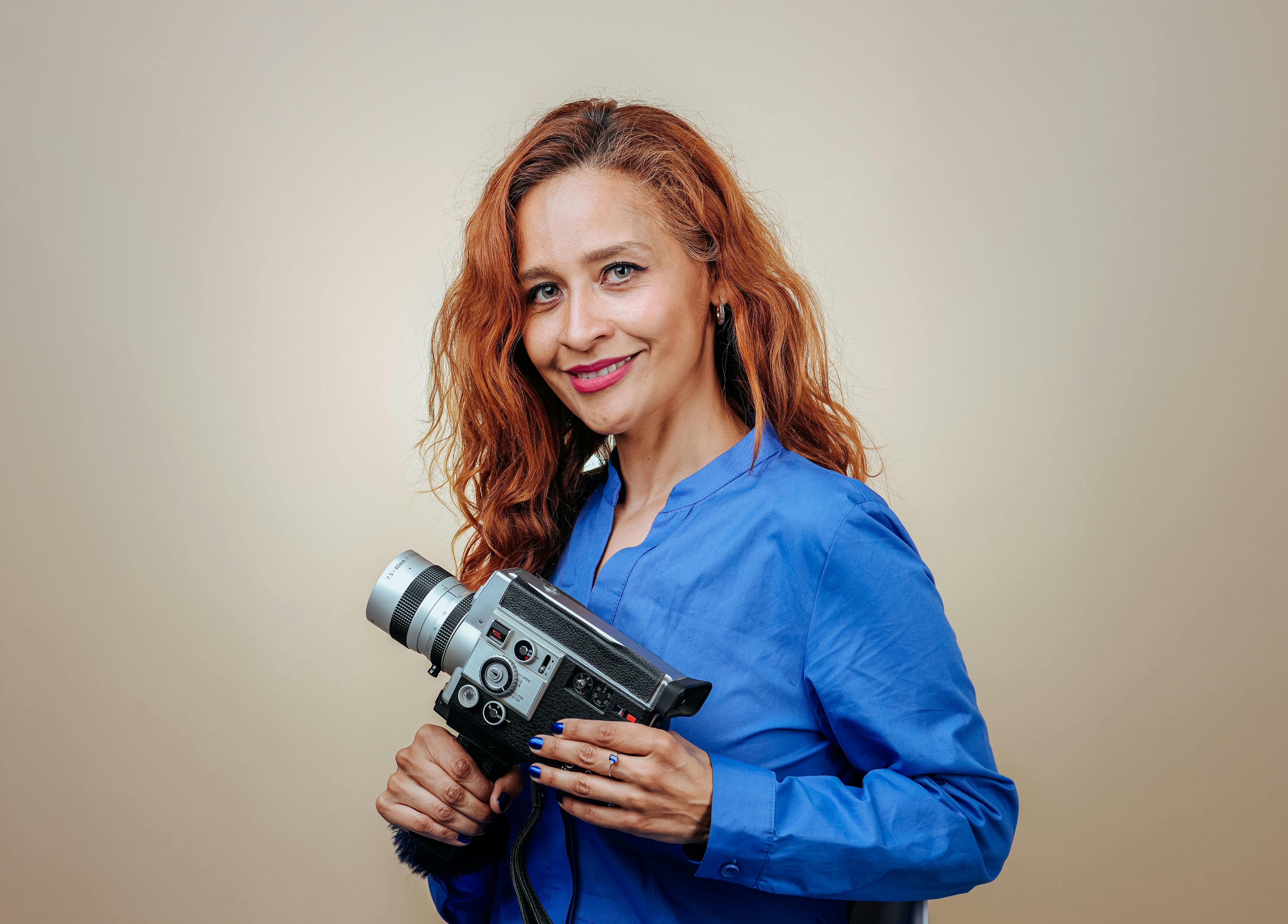 Smiling woman with red hair holds a vintage camera in a studio setting.