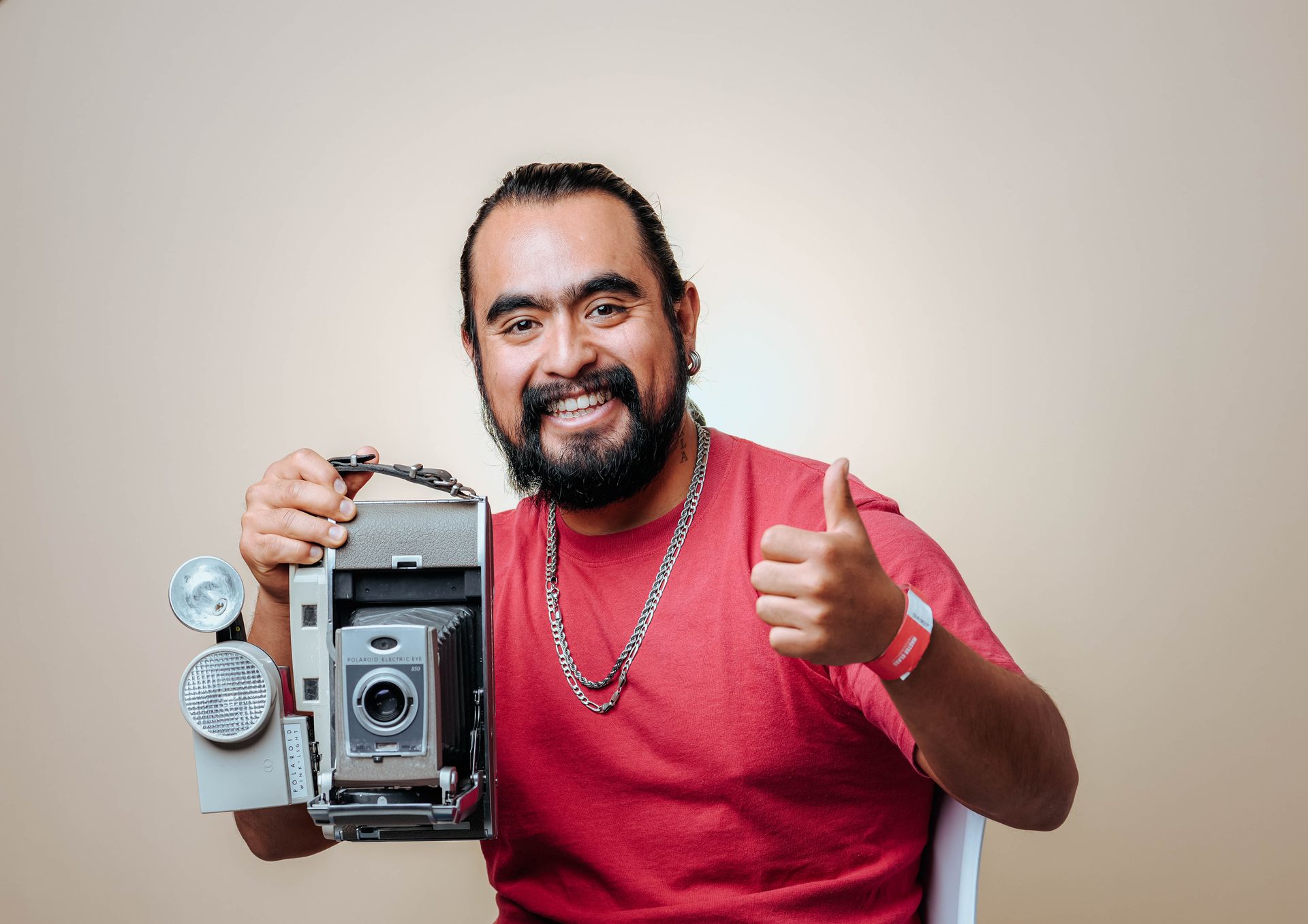 Smiling man with a beard gives a thumbs up while holding a vintage camera in a studio setting.