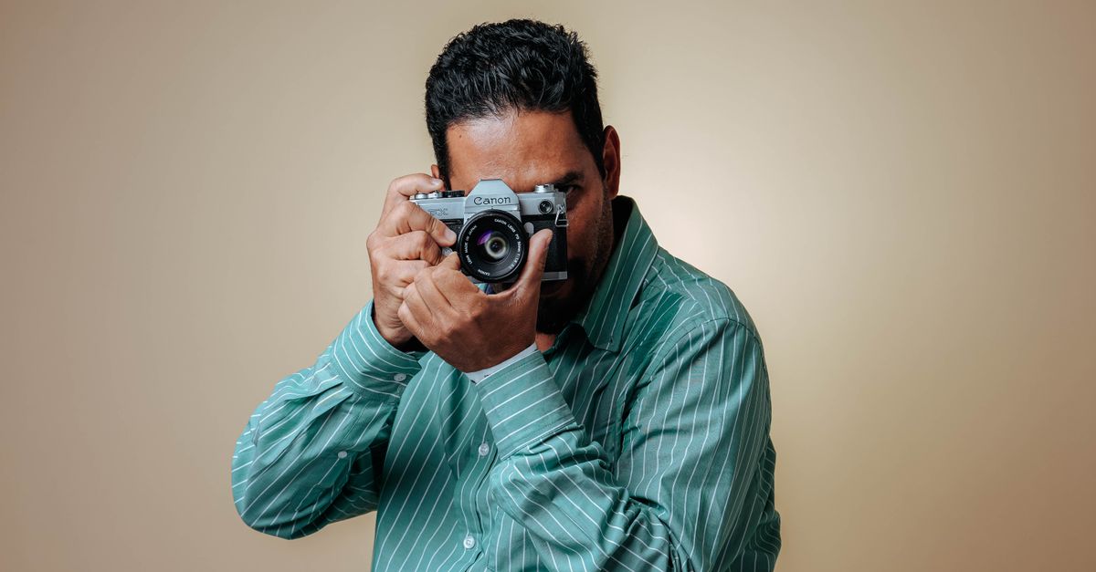 Man posing with a vintage camera in a studio setting wearing a striped green shirt.