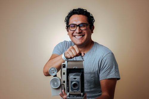 Free Man in glasses holding a vintage camera, smiling in a studio shot with a neutral background. Stock Photo