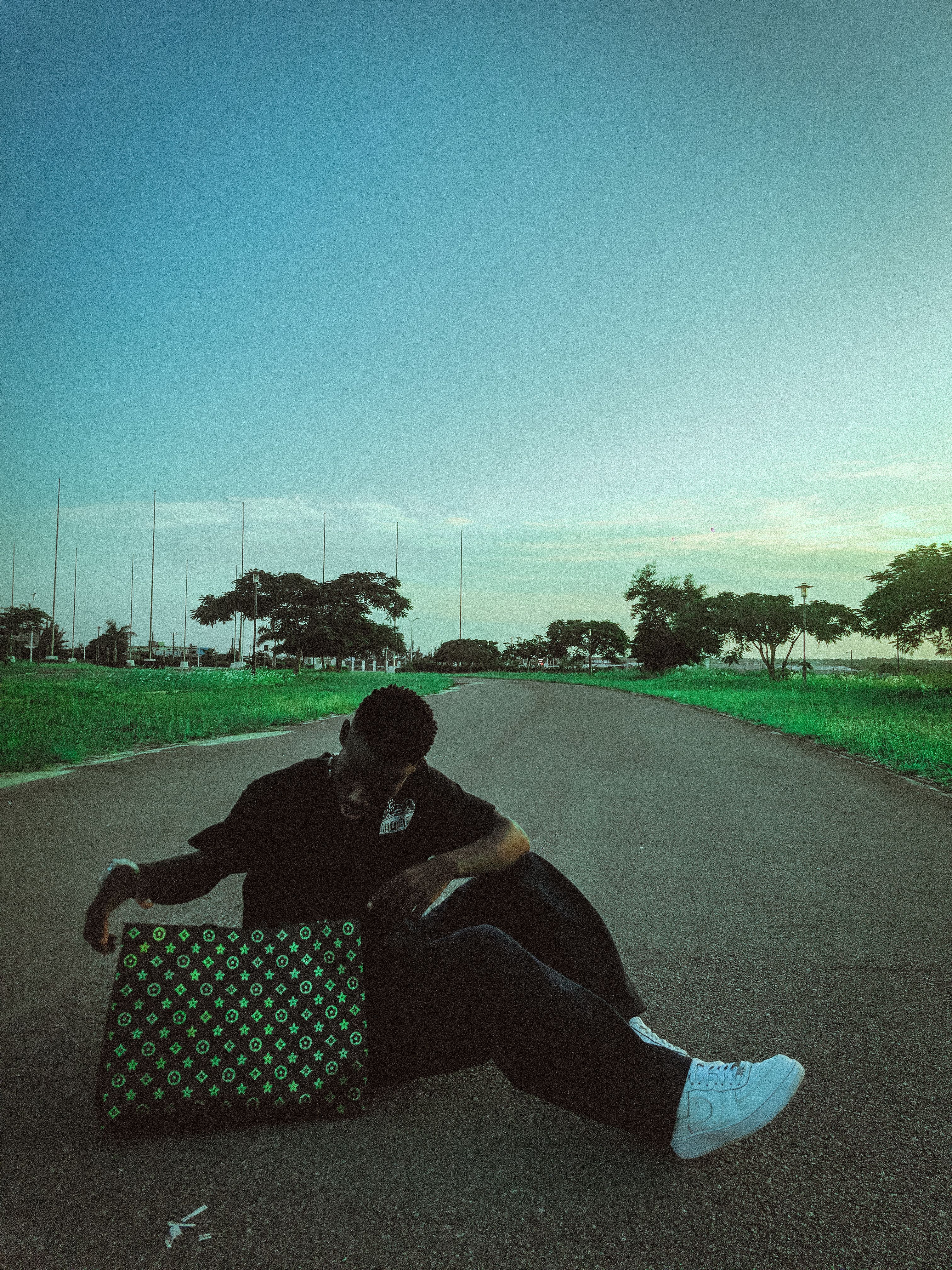 A man sits stylishly on a road holding a designer bag under a clear blue sky, exuding urban cool.