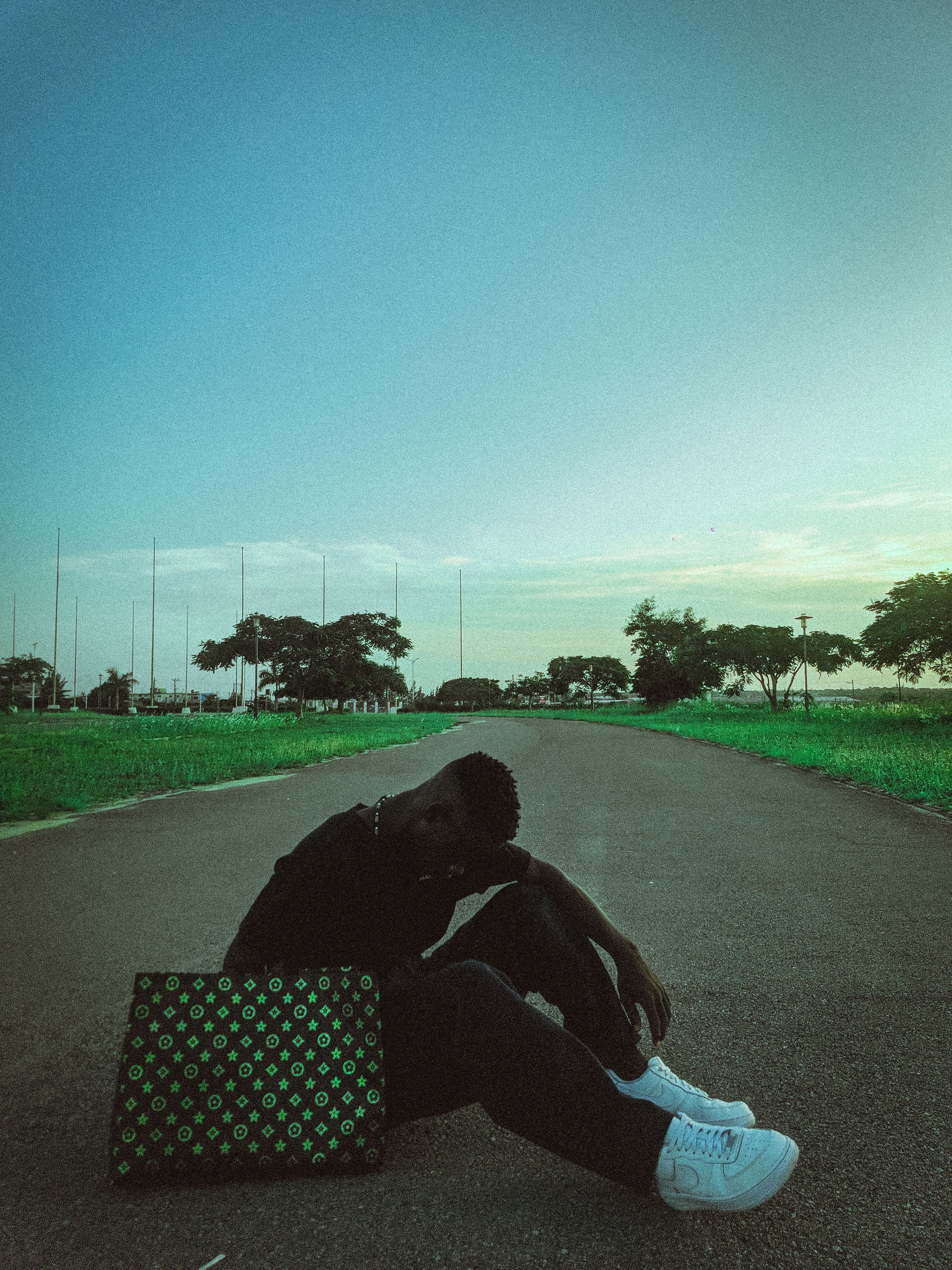 A young man sits contemplatively on a suburban road, bag by his side, during daytime.