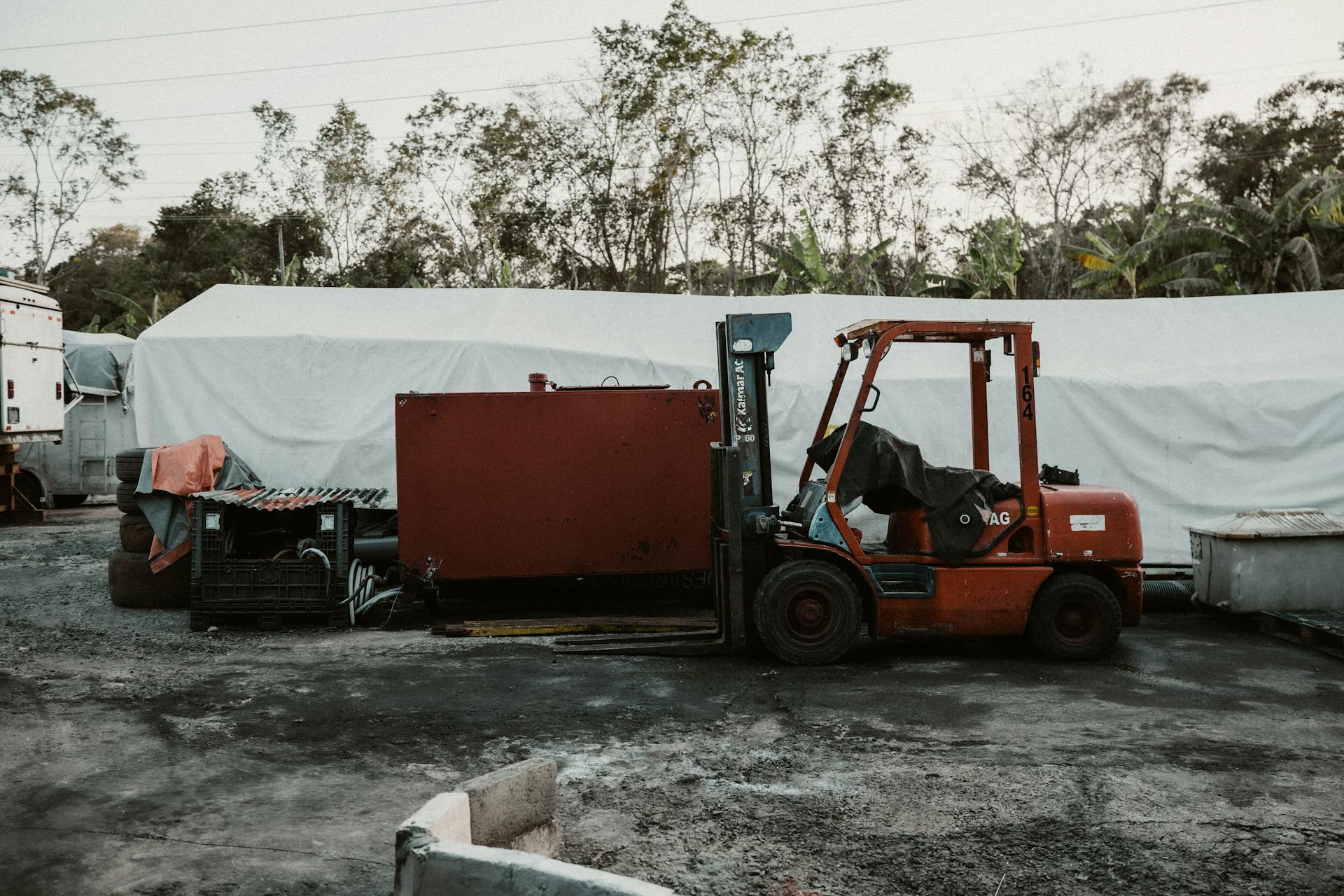 A red forklift parked in a rural area with equipment and trees in the background.