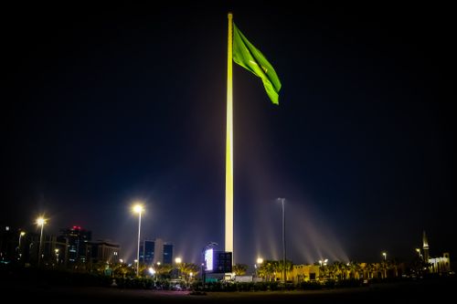Free Majestic Saudi Arabian flag illuminated against the night sky, surrounded by cityscape lights in Riyadh. Stock Photo