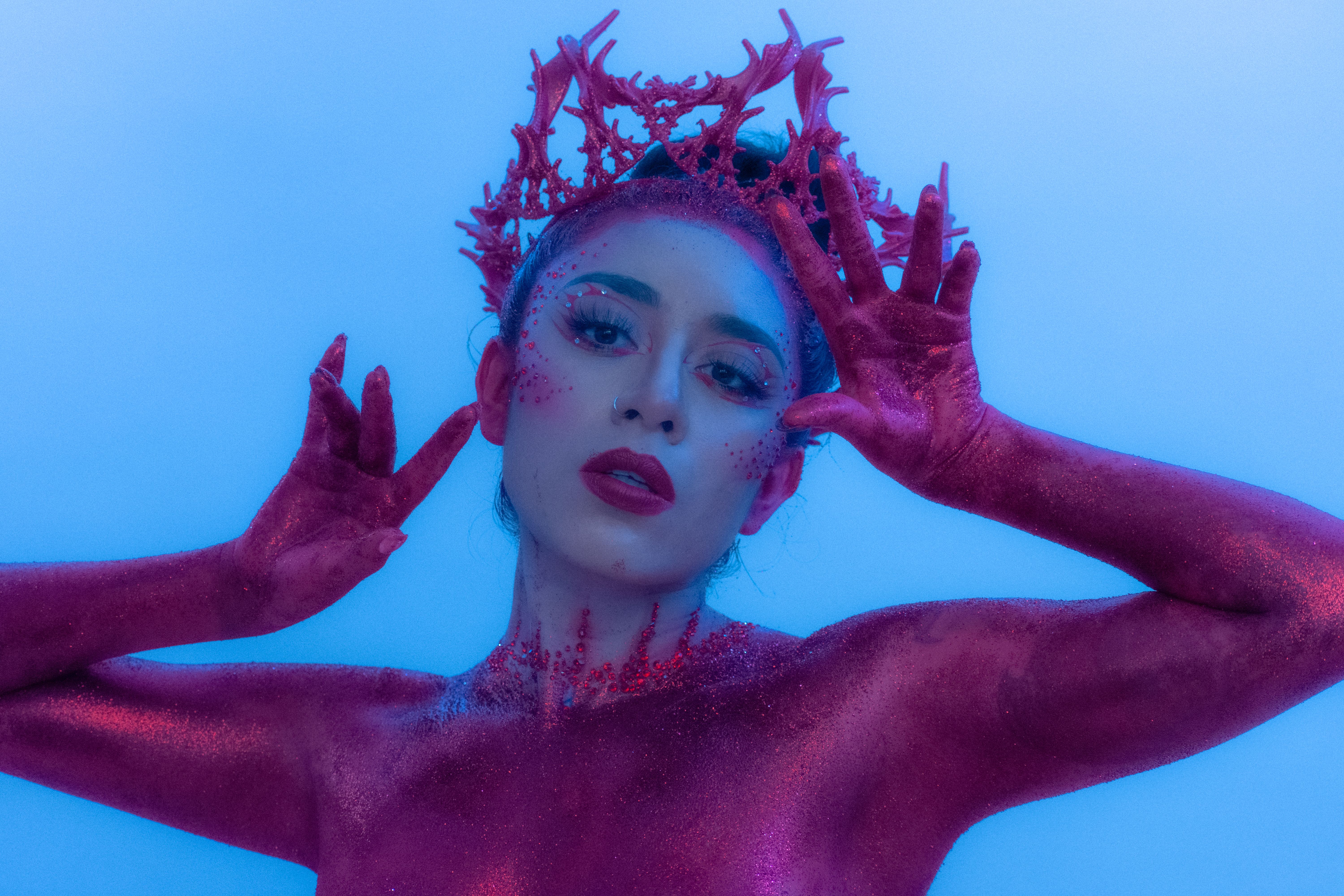 A stunning artistic portrait of a woman adorned in shimmering pink body paint and a coral crown, under blue lighting.