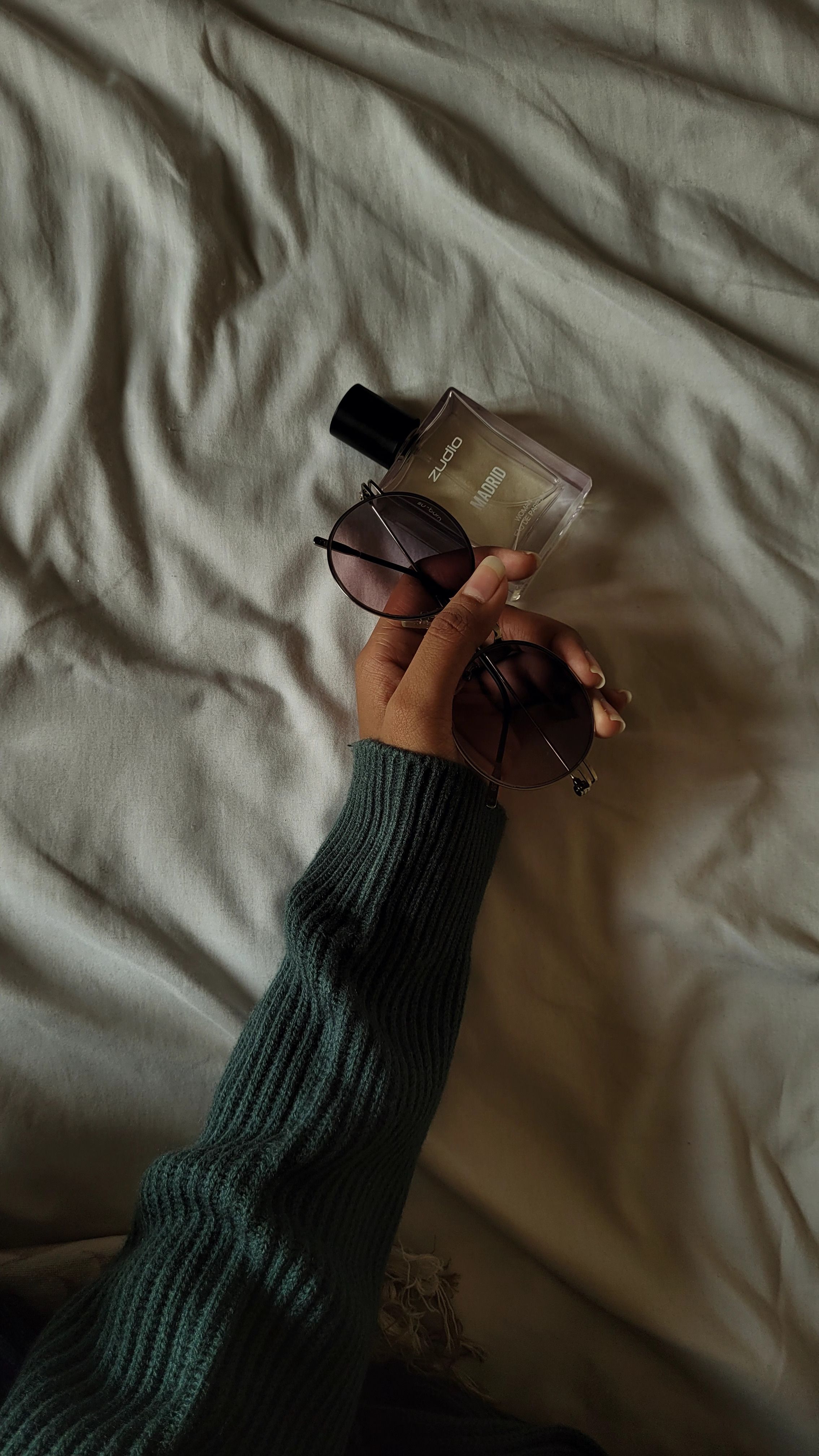 Close-up of a hand holding a perfume bottle and sunglasses on a textured fabric.