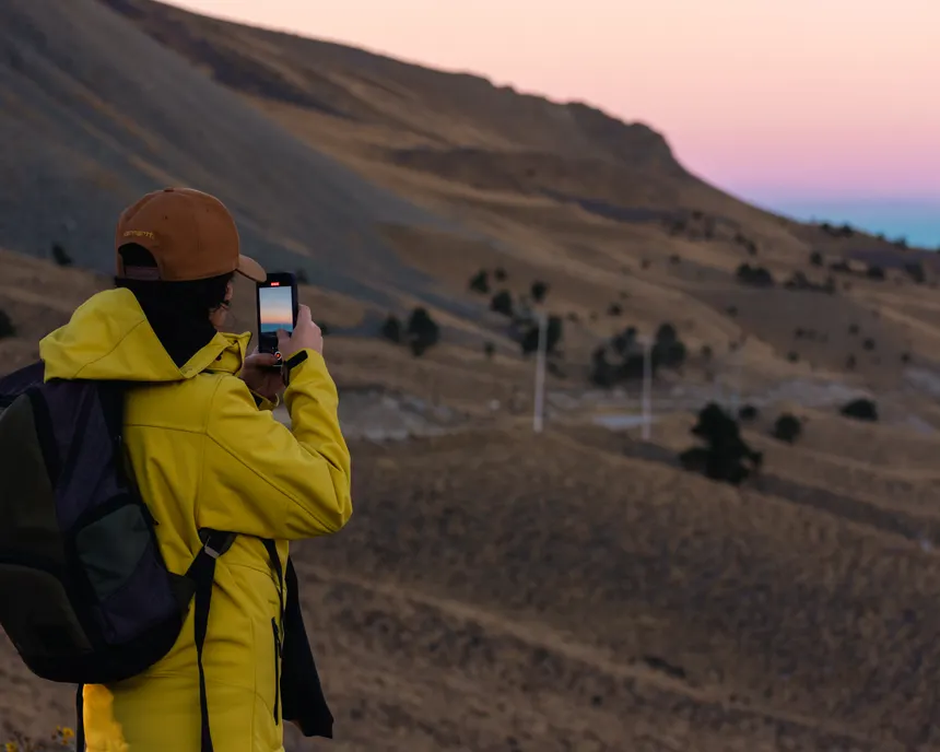 A woman in yellow jacket photographs a picturesque mountain sunset outdoors.
