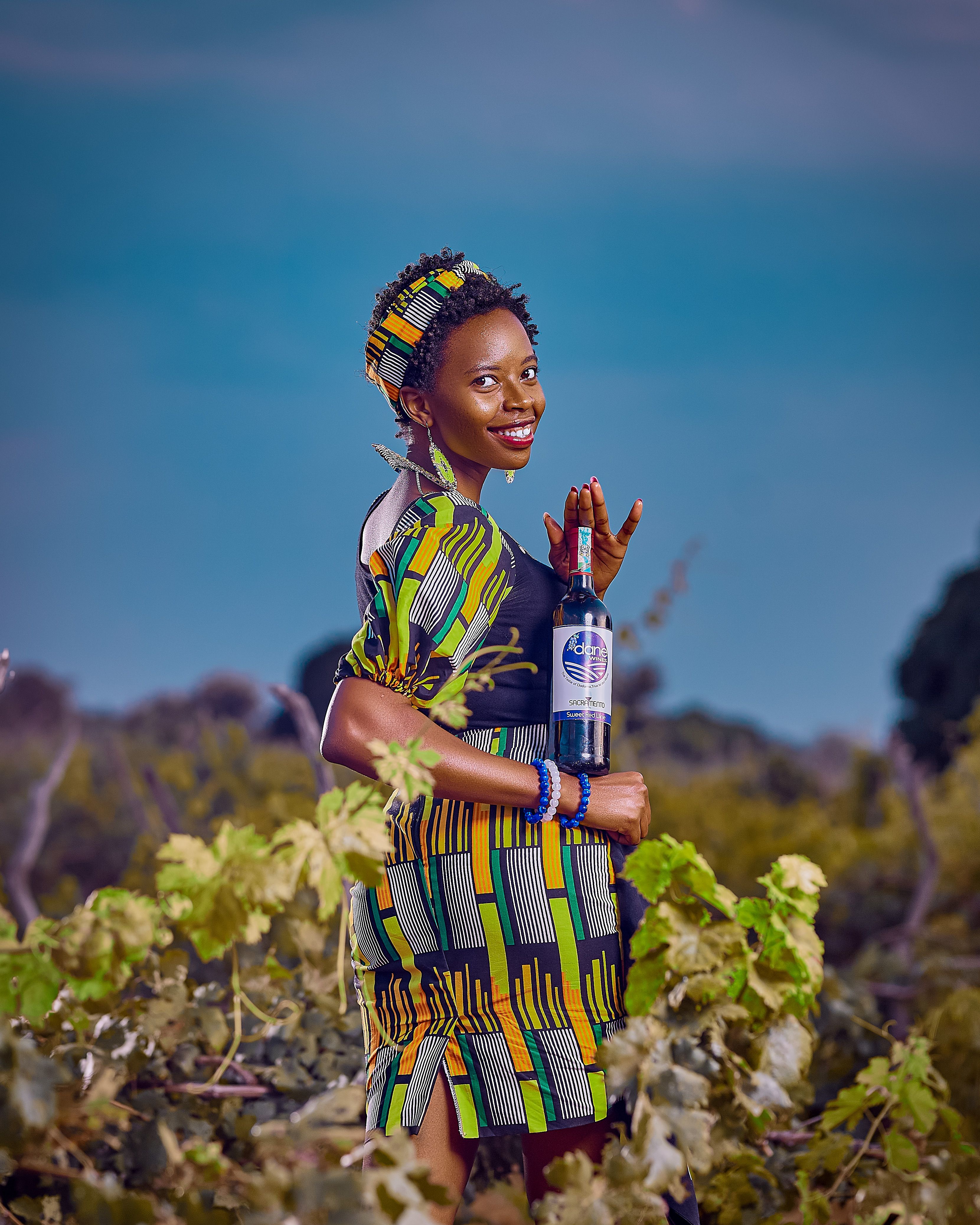 Smiling woman in traditional attire holding a wine bottle in a lush vineyard setting.