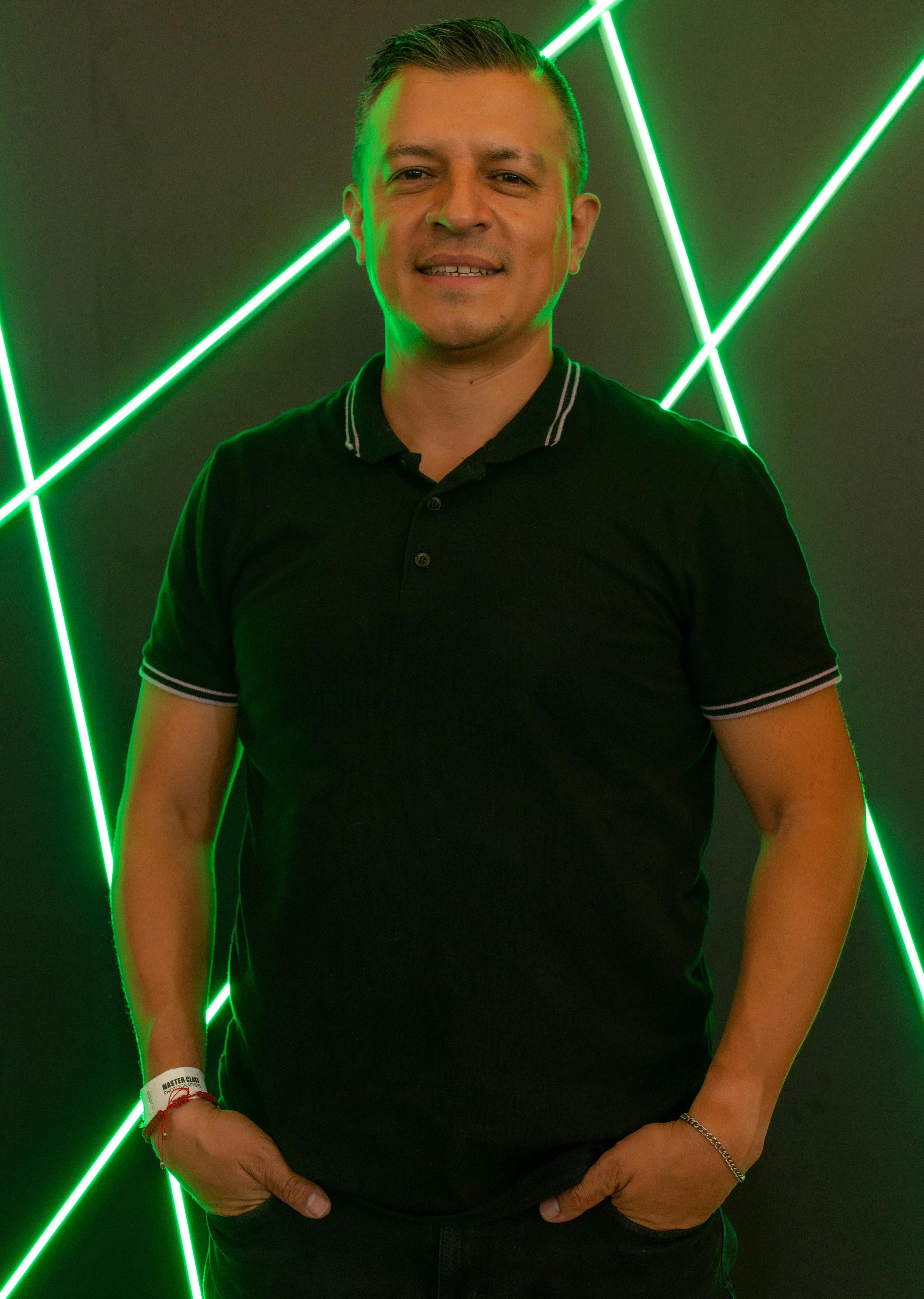 Man smiling and posing with hands in pockets, set against a vibrant green neon line background in a studio.