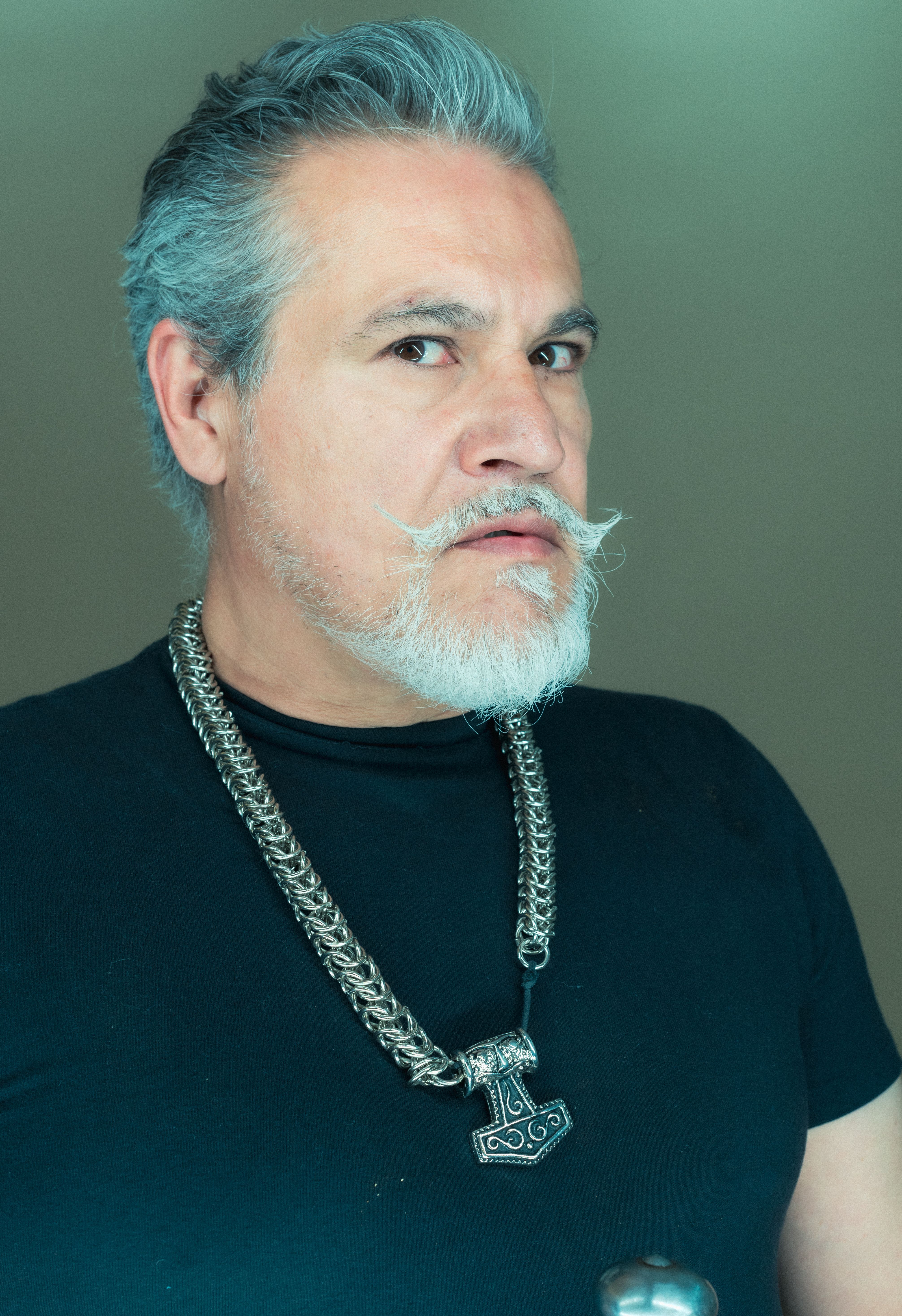 Portrait of an older man with a waxed mustache, gray hair, and a silver necklace, taken in a studio setting.