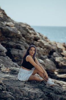 A woman with dyed hair sitting on rocky shore, gazing towards the ocean under daylight.