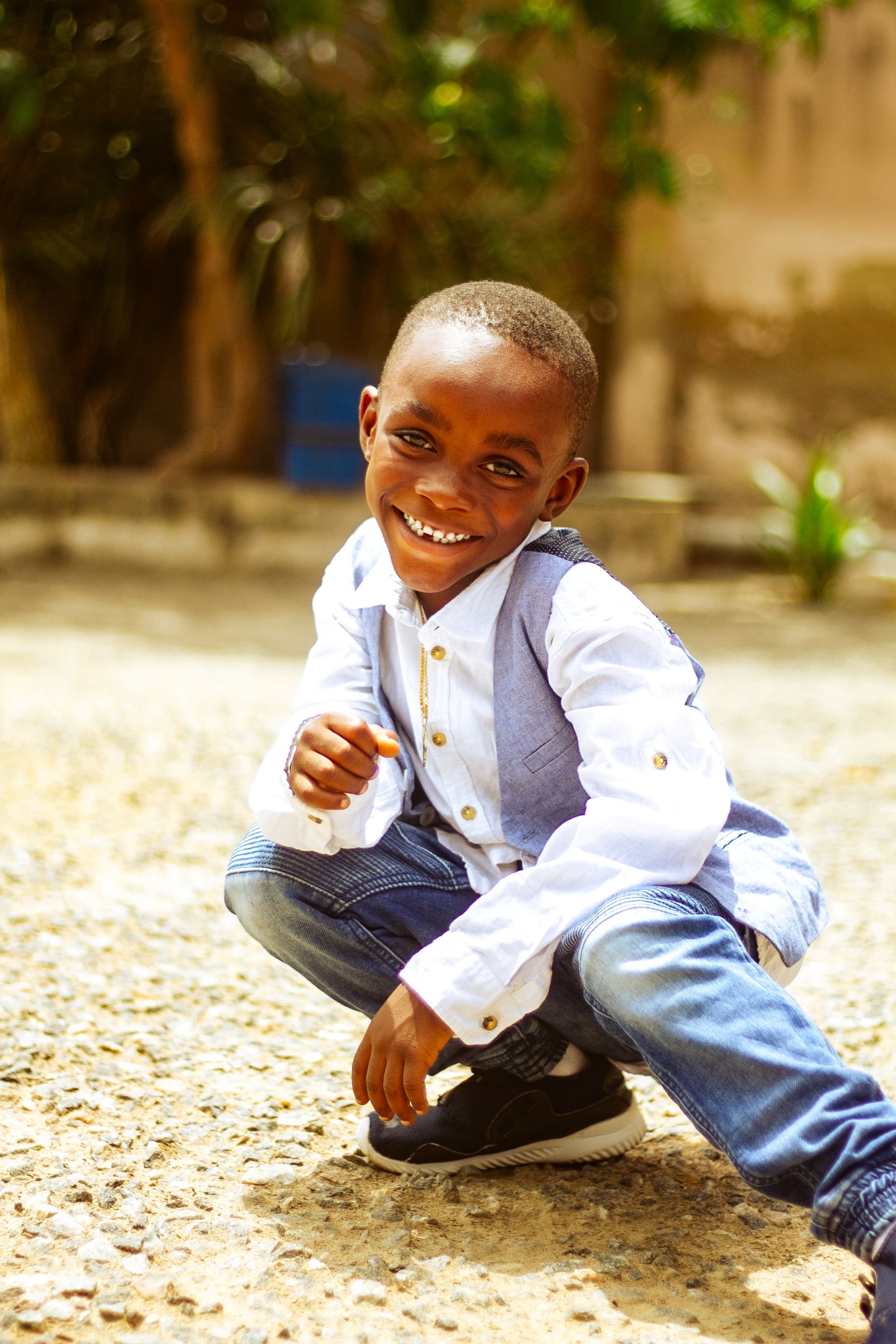 A cheerful boy in casual attire squatting outdoors and smiling brightly on a sunny day.