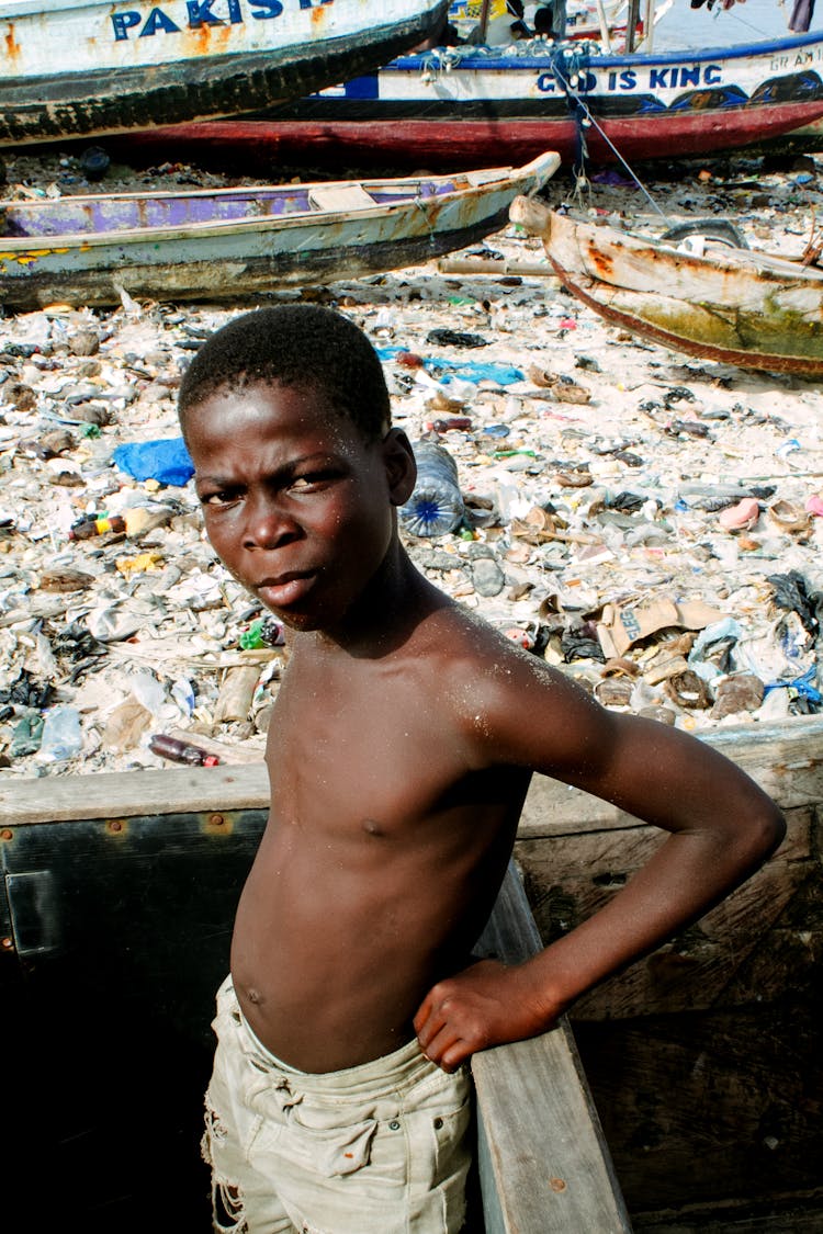 Young Boy On Boat Sailing Through Polluted River Covered In Trash