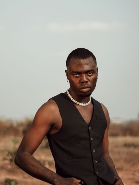 Elegant portrait of a man wearing a black waistcoat and pearls, set against an outdoor backdrop.