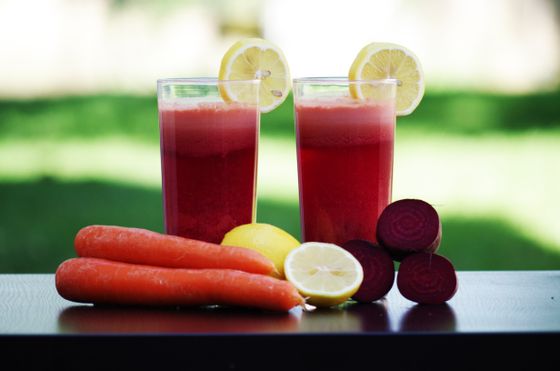 Two glasses of deep red beet juice with lemon slices beside carrots, lemons and beets
