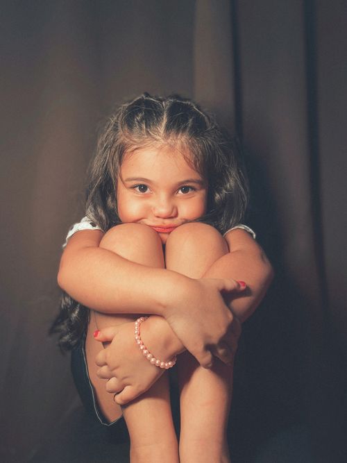 Free Cute young girl sitting with knees up, smiling warmly indoors. Stock Photo