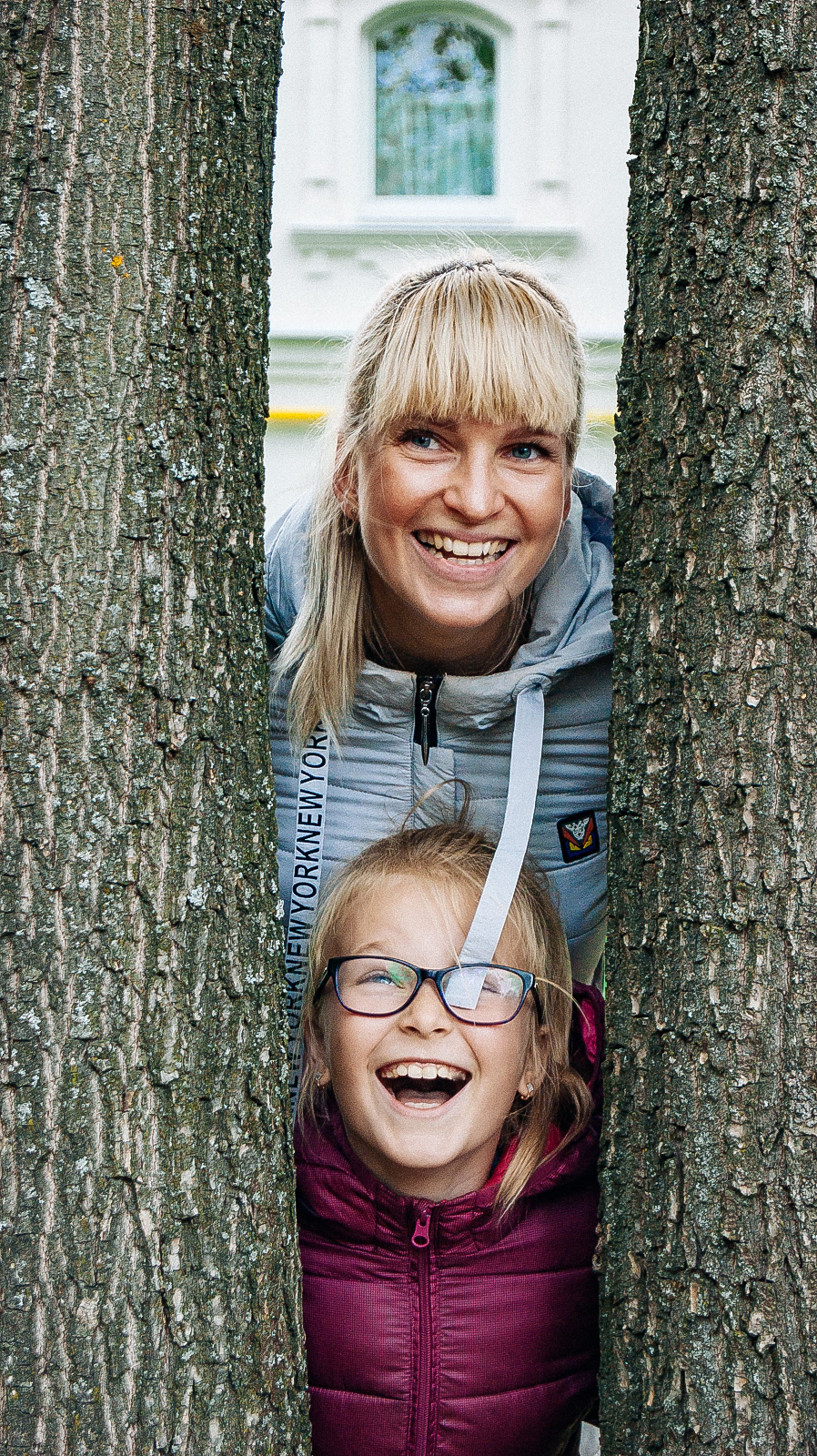 Happy mother and daughter smiling while posing between tree trunks outdoors.
