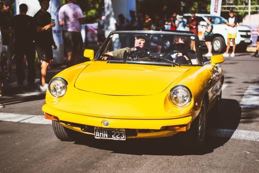 A classic yellow Alfa Romeo spider convertible driving at a lively urban event with onlookers.