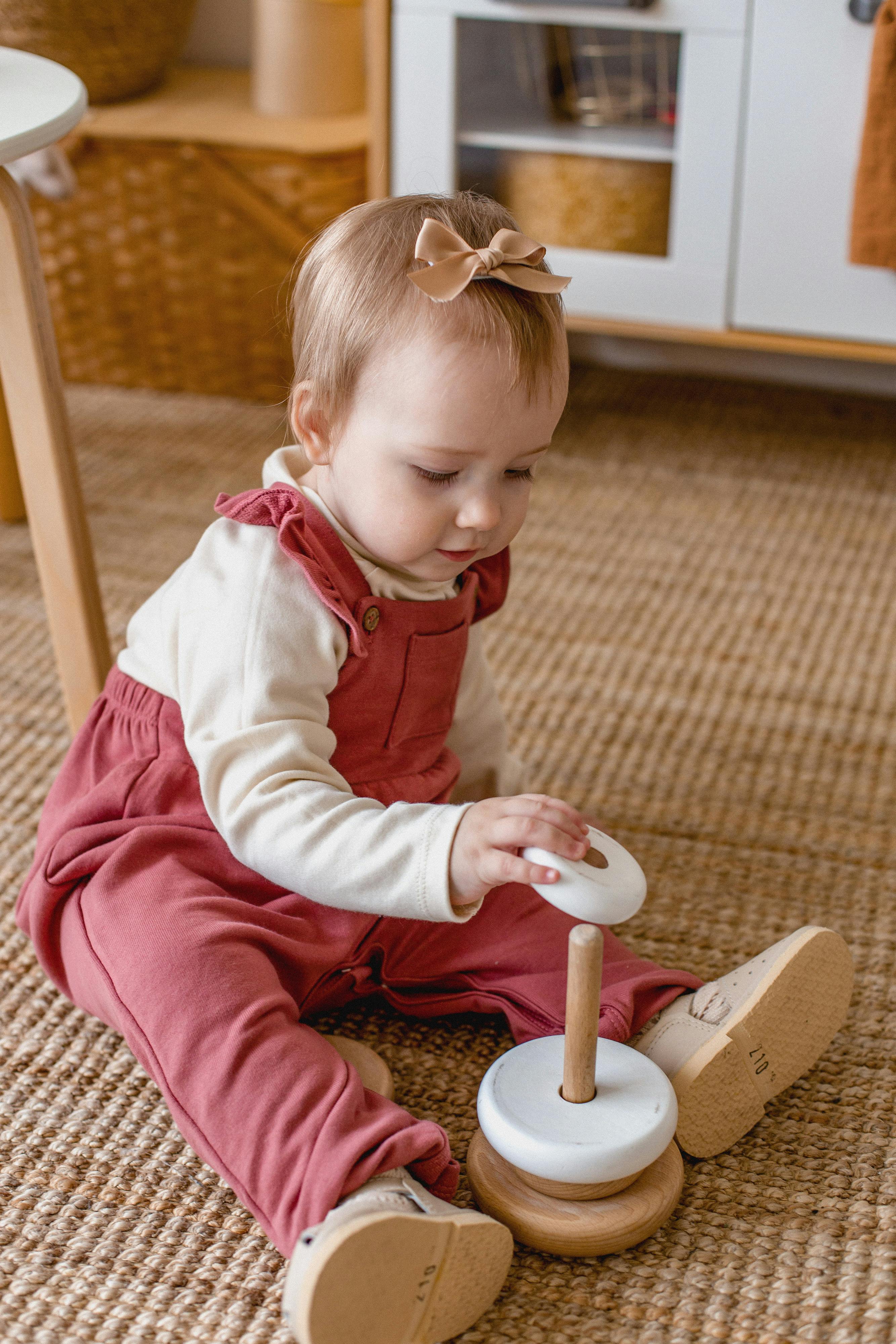 Cute baby in overalls playing with a wooden toy on a carpeted floor indoors.
