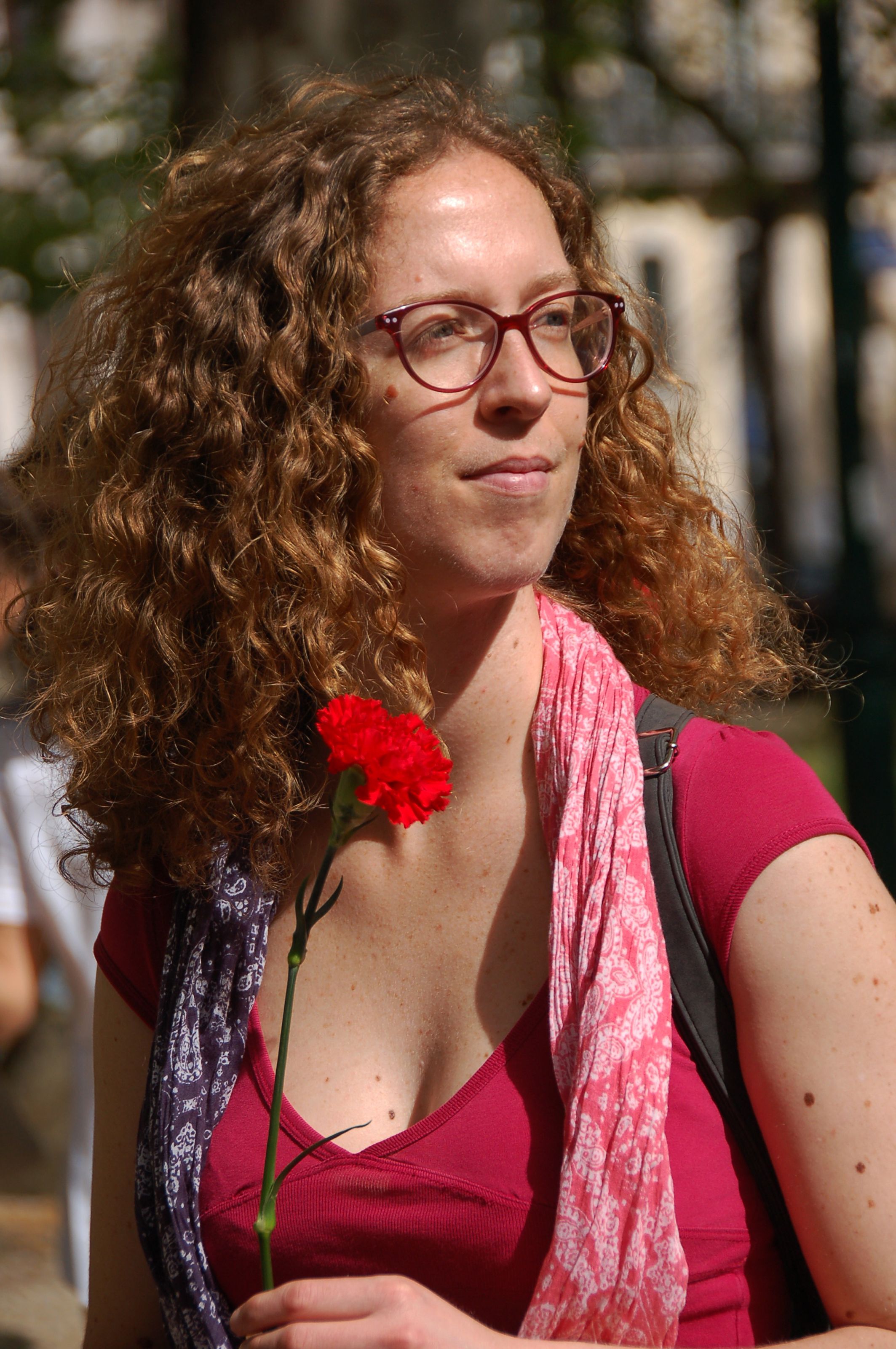 Portrait of a woman with curly hair and eyeglasses holding a red carnation in a sunlit Lisbon park.