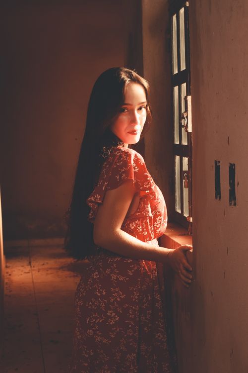 Free A woman in a red dress stands by a sunlit window, creating a warm and serene atmosphere. Stock Photo