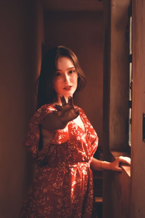 Free A woman in a red dress reaches out while standing by a sunlit window. Stock Photo