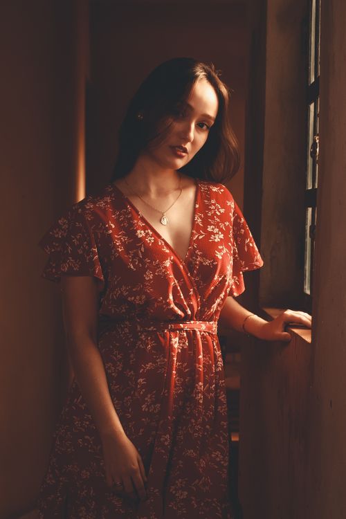 Free A beautiful woman stands by a window wearing a red floral dress, bathed in warm light. Stock Photo