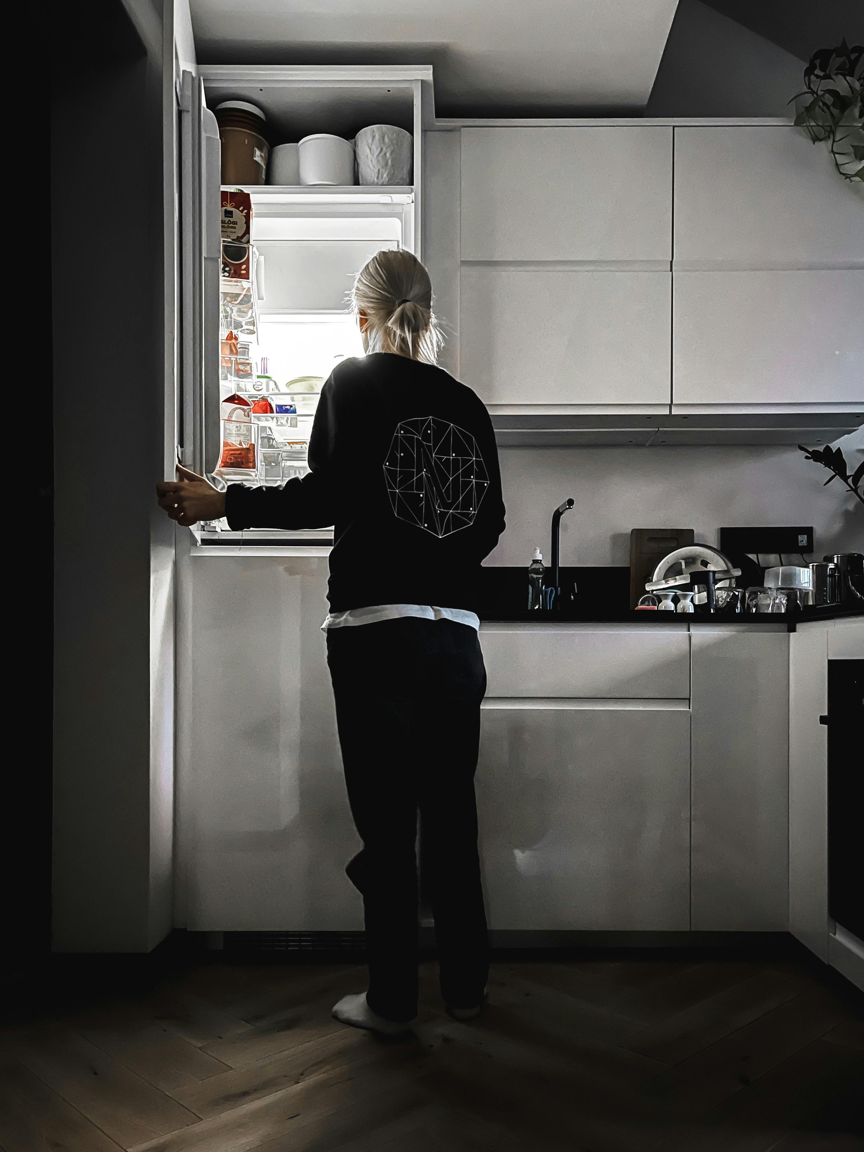 Free Adult woman in a kitchen looking into an open refrigerator. Back view. Stock Photo