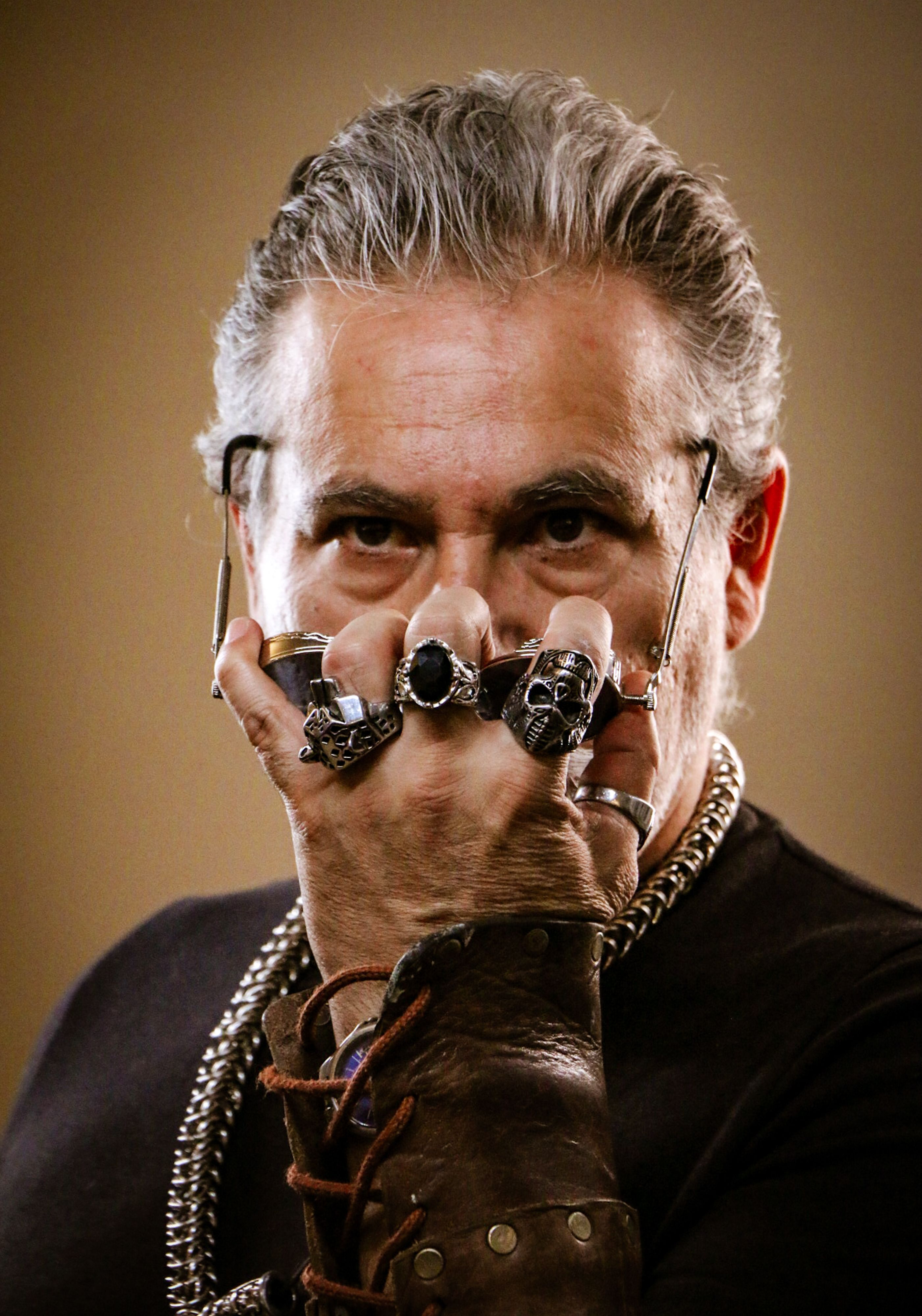 Portrait of a stylish man with gray hair, wearing multiple rings and glasses against a warm backdrop.