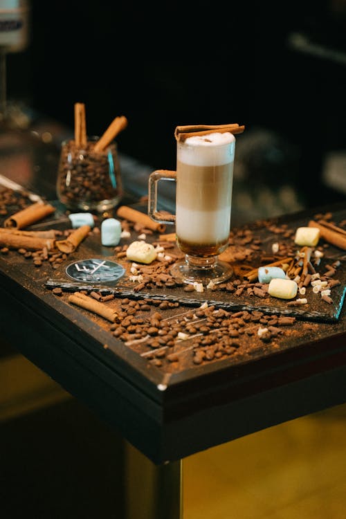 A Glass with Latte Macchiato Standing on a Table with Scattered Coffee Beans and Cinnamon Sticks