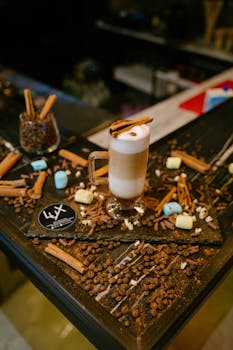 Latte macchiato topped with cinnamon stick surrounded by coffee beans and marshmallows on a table.