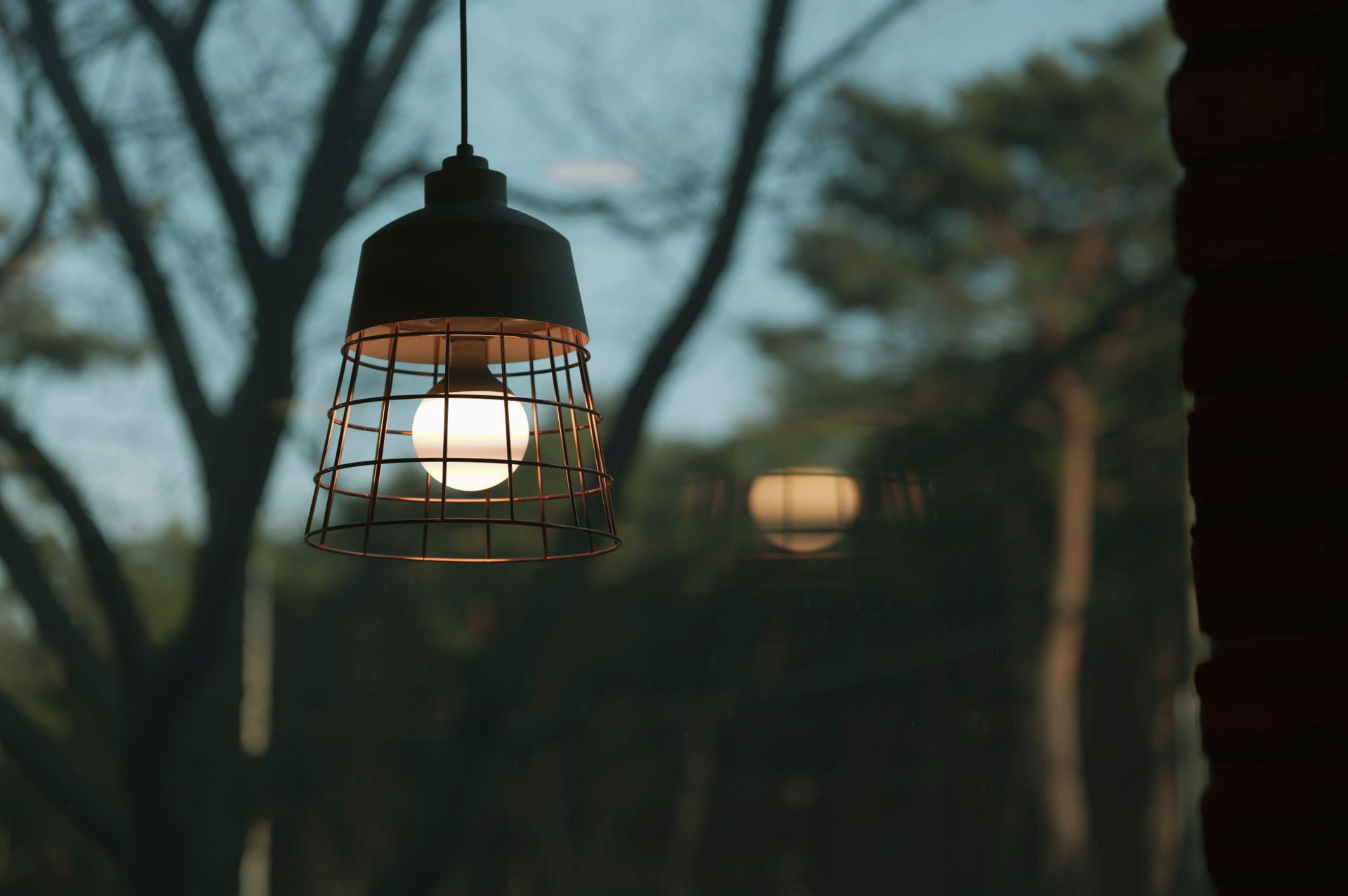 A close-up of a cage pendant light casting a warm glow, set against a blurred outdoor background with trees.
