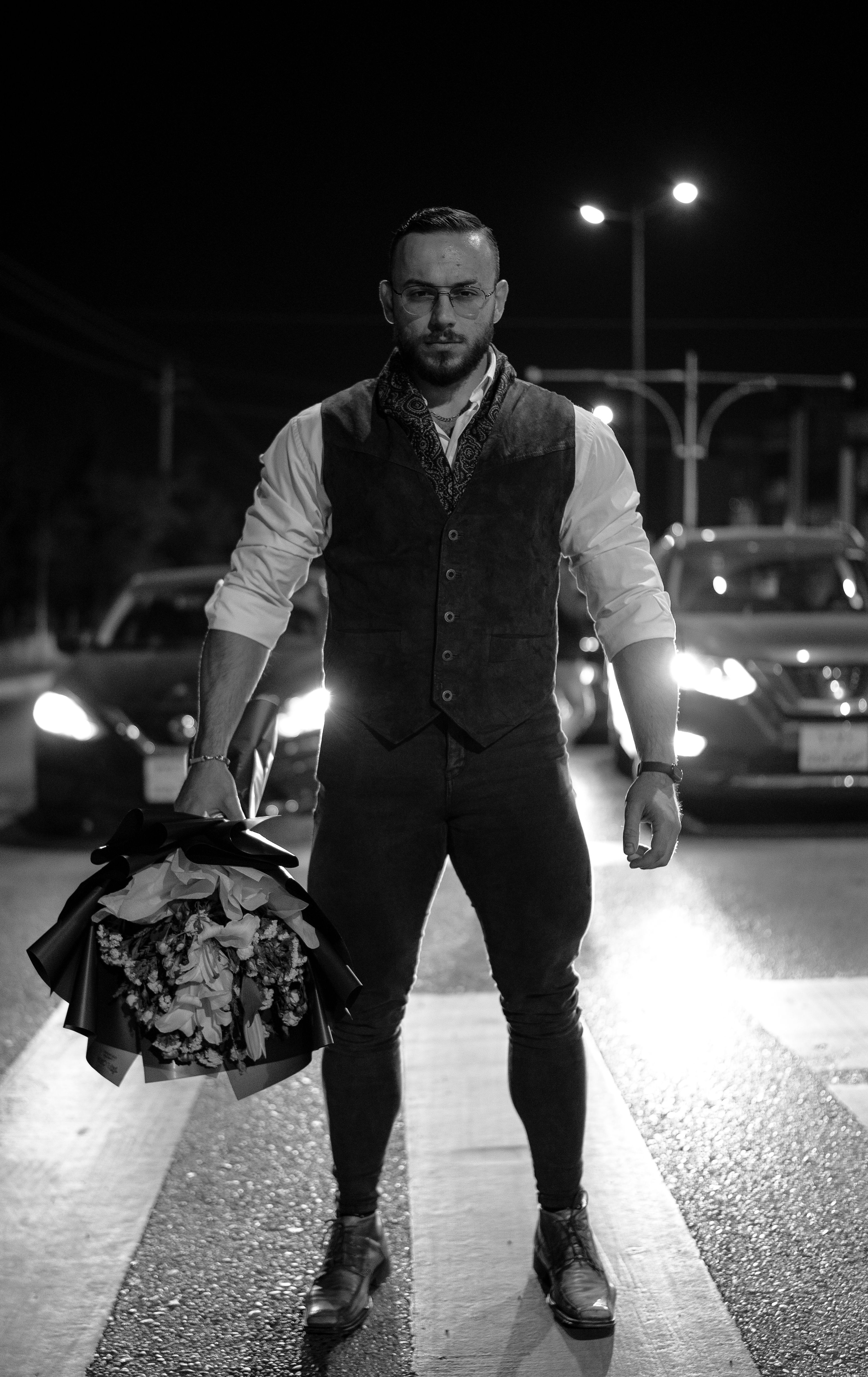 Fashionable man in Sulaymaniyah, Iraq, posing at night on city street holding flowers.