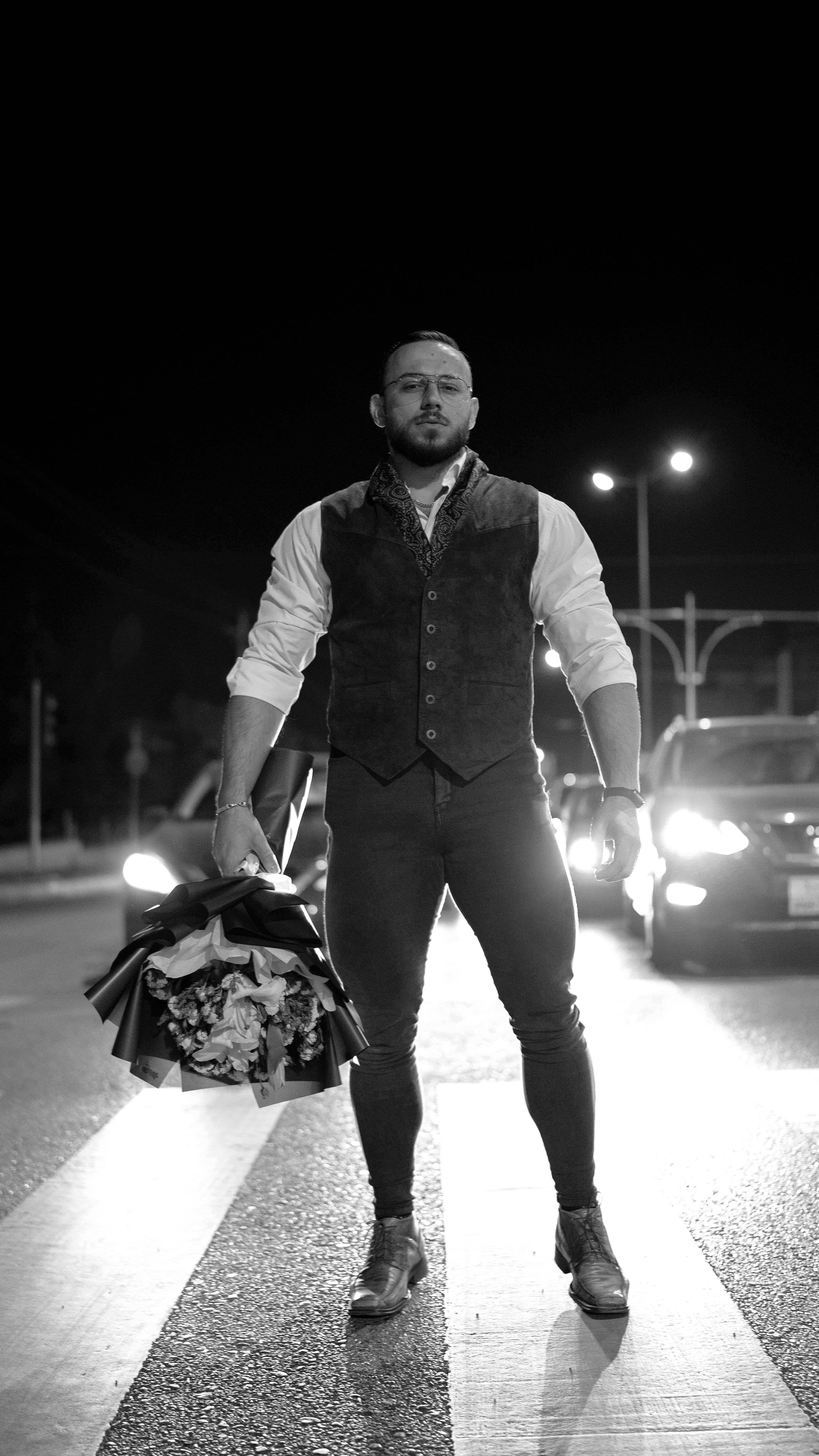 A man in a fashionable vest holds a bouquet on a city street at night.