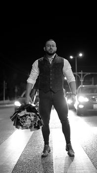 A man in a fashionable vest holds a bouquet on a city street at night.