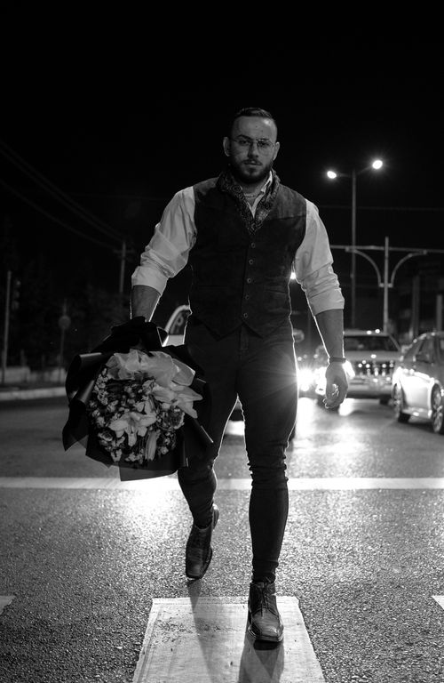 Free A fashionable man in a vest carries a bouquet while standing on an urban street at night in Sulaymaniyah. Stock Photo