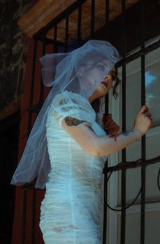 Elegant bride stands by window bars in a vintage wedding dress, exuding mystery.