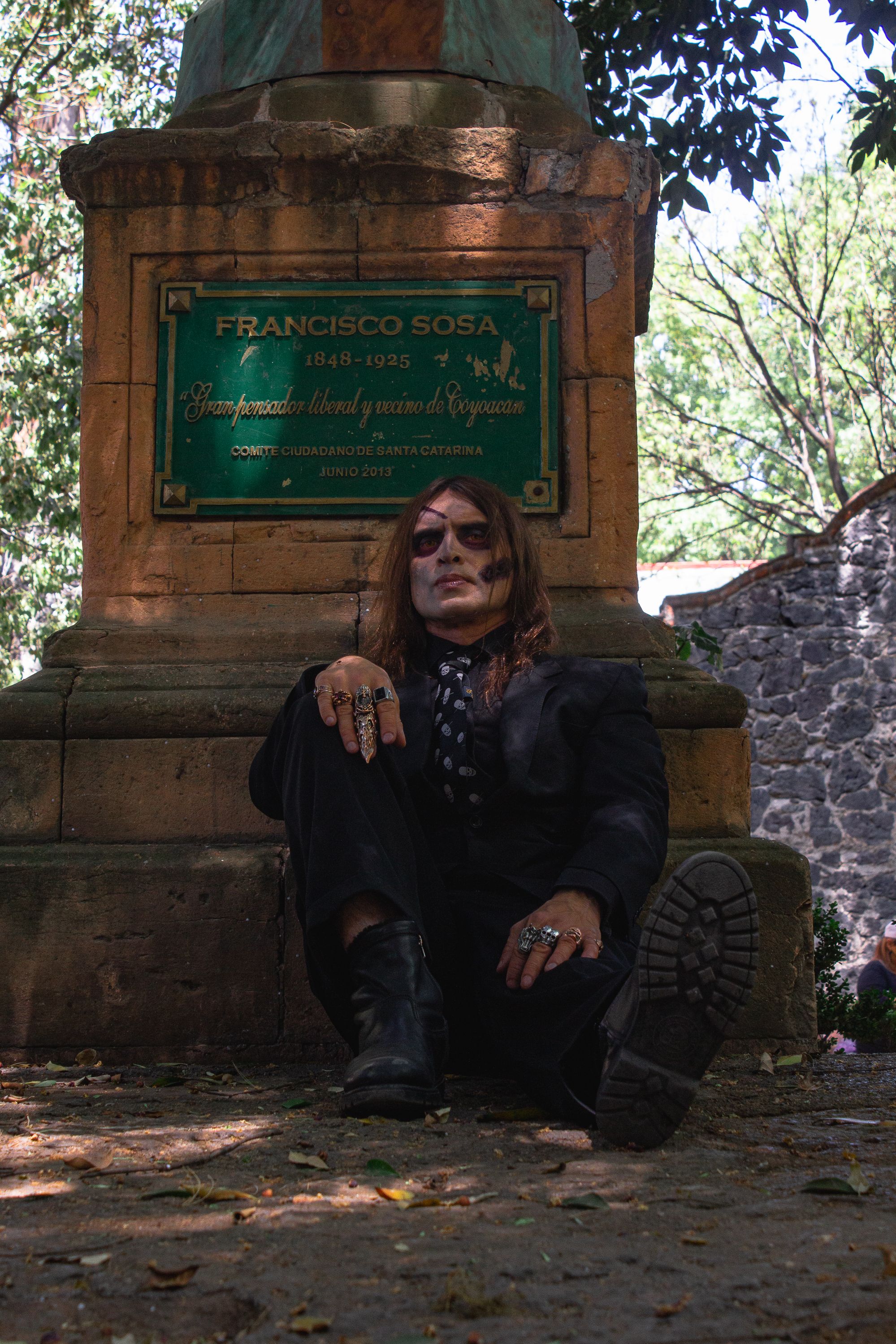 Portrait of a man in gothic attire sitting by a stone monument outdoors.