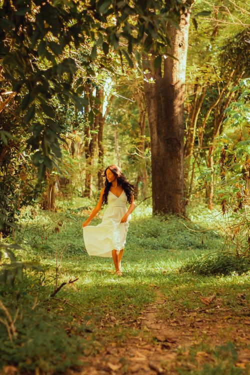 Free A woman in a flowing white dress strolls gracefully through a sun-dappled forest pathway. Stock Photo