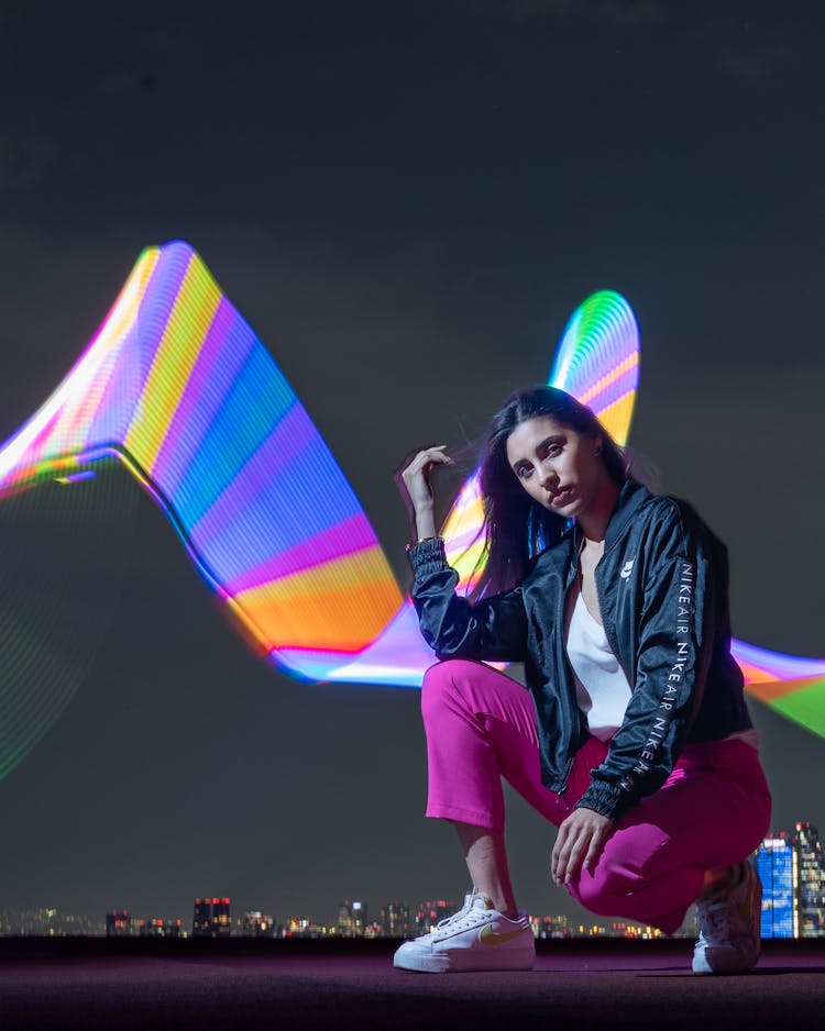 Portrait Of A Pretty Brunette Crouching In Front Of A Colorful Light Painting At Night