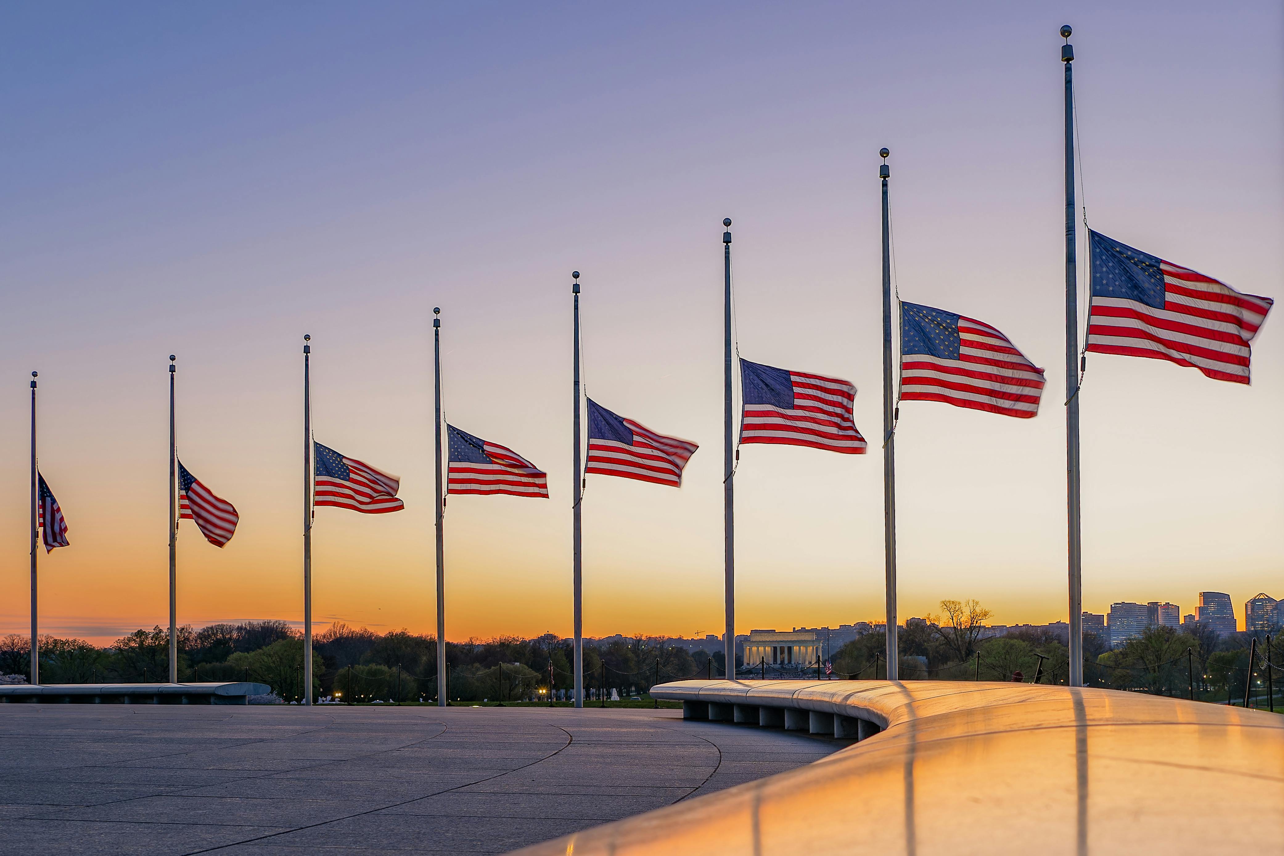 American flags at half-mast surround the Washington Monument during a vibrant sunset.