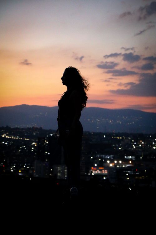 Free A woman stands silhouetted against a vibrant sunset with city lights below, capturing a serene urban moment. Stock Photo