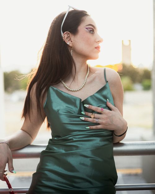 Free Stylish female model in an elegant green dress with a chic pose and cool accessories against a sunset backdrop. Stock Photo