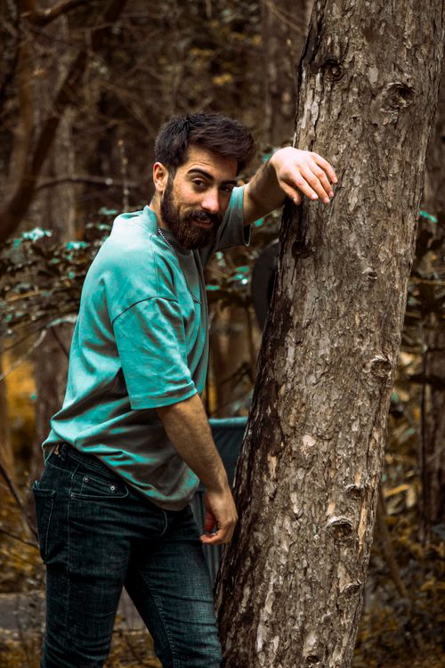 Free Man with a beard leaning against a tree in a lush forest, wearing casual clothing. Stock Photo