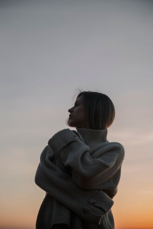 Free A woman in a warm sweater stands silhouetted against a sunset sky, evoking a calm and reflective mood. Stock Photo