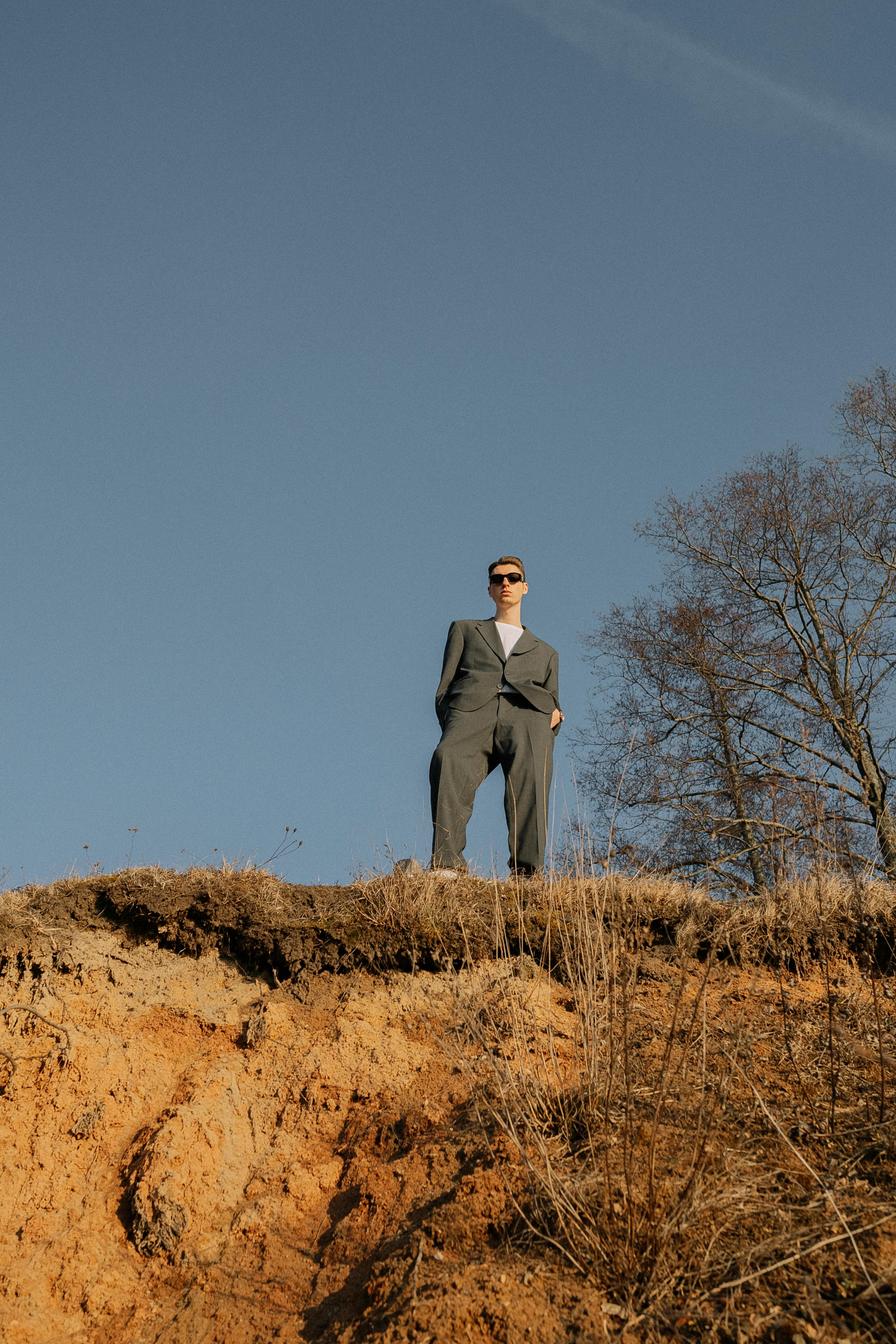 A man in a suit stands on a hilltop under a clear sky, exuding confidence and style.