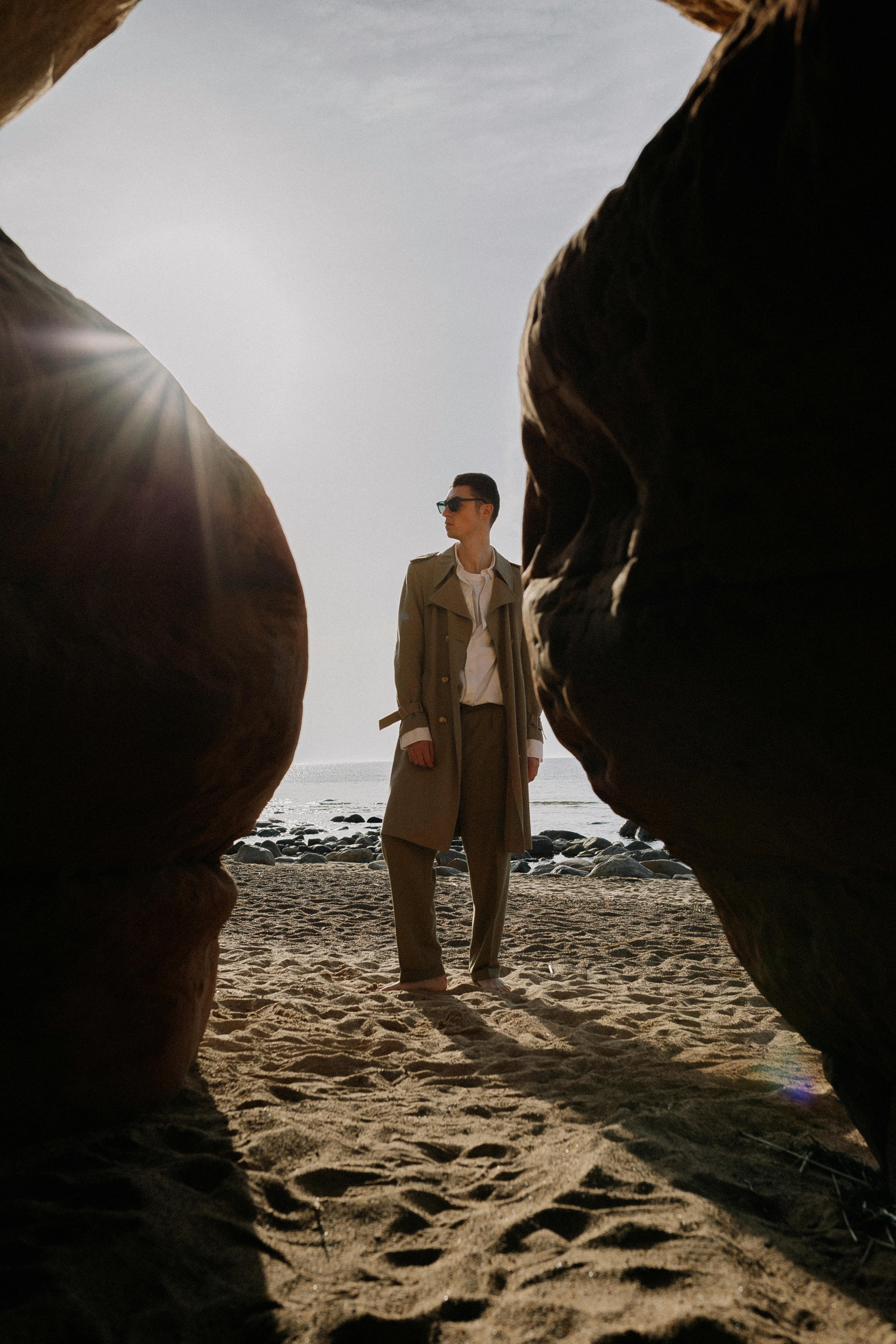 A fashionable person stands between rocky formations on a sandy beach under the sun.