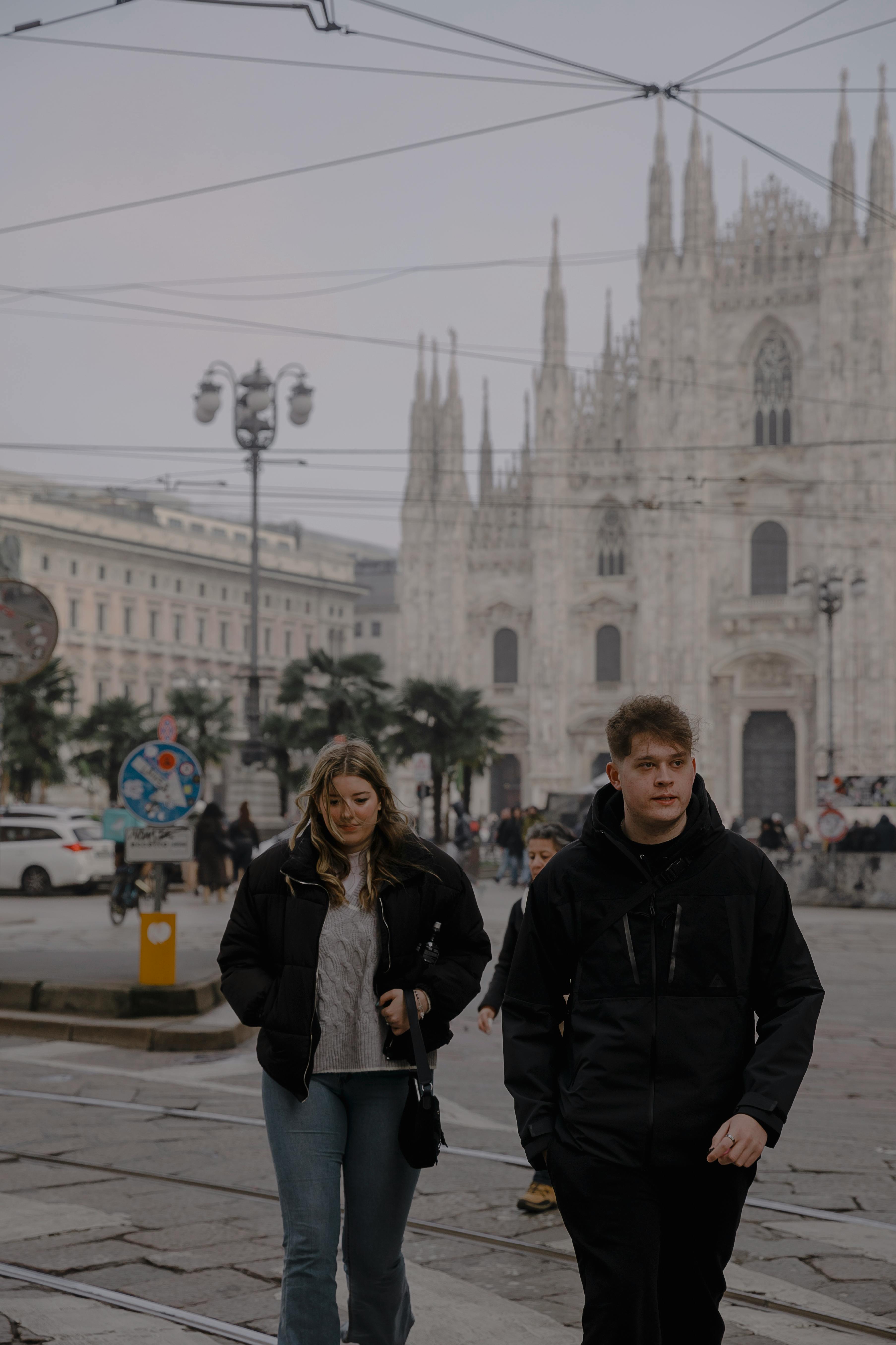 Pedestrians walking near the iconic gothic architecture of Milan Cathedral on a cloudy day, capturing urban life.