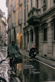 A cyclist in a raincoat rides through wet, historic city streets, reflecting a city life in the rain.