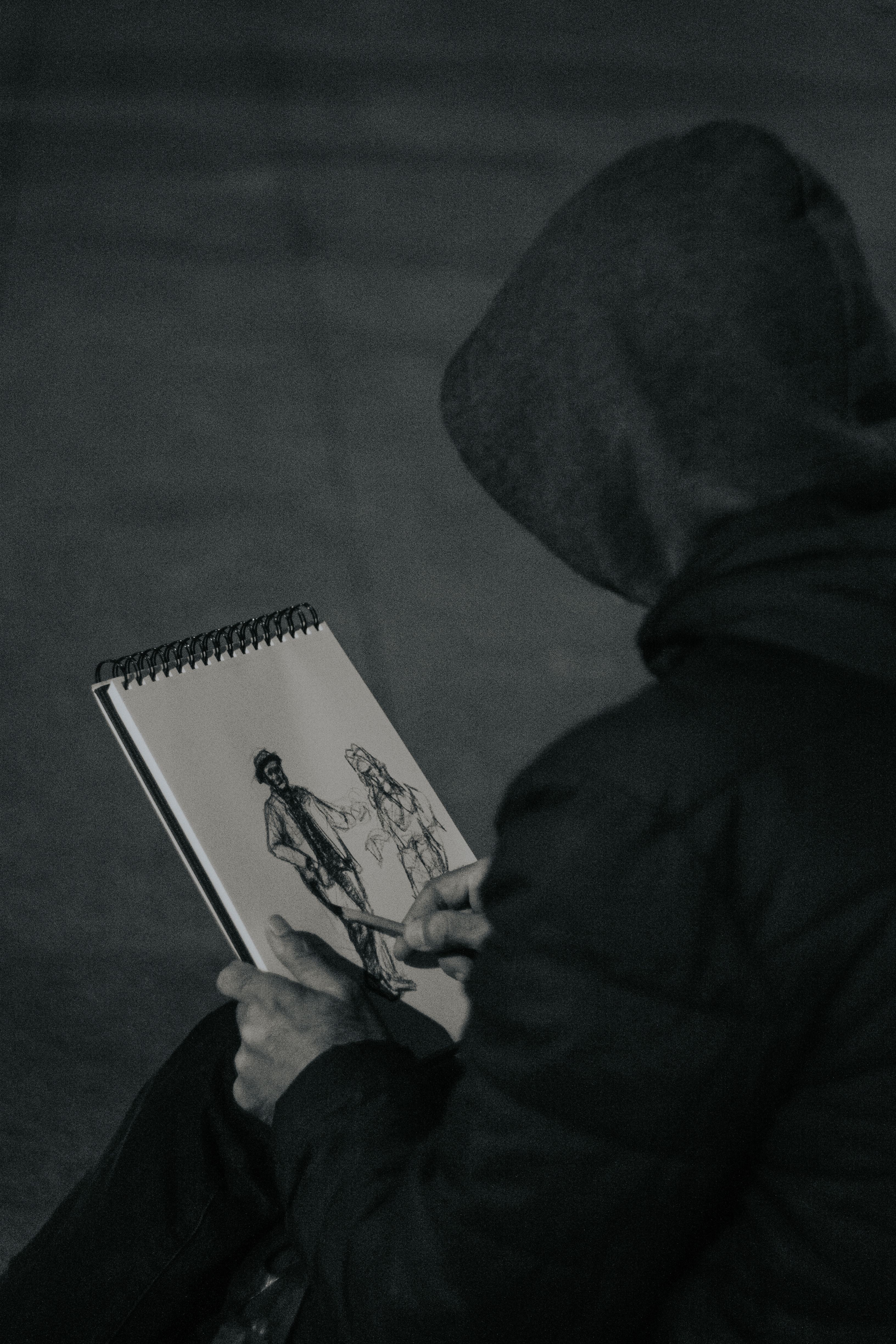 A person in a hood draws in a sketchbook in dim lighting, showcasing artistic talent.
